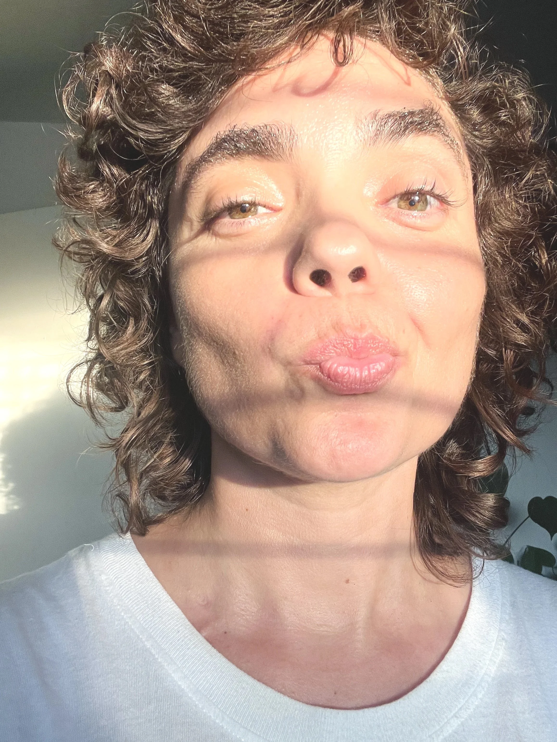 Close-up of a person with curly hair, light skin, and wearing a white top, looking upward in natural light.