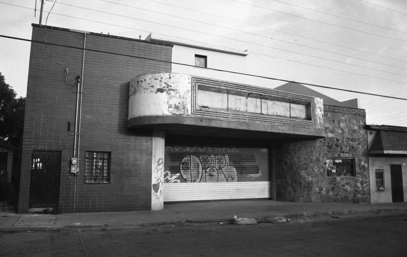 An old, abandoned building with graffiti on a rolling shutter, peeling paint, and a small window with bars, featuring brick, concrete, and stone construction with wires overhead.