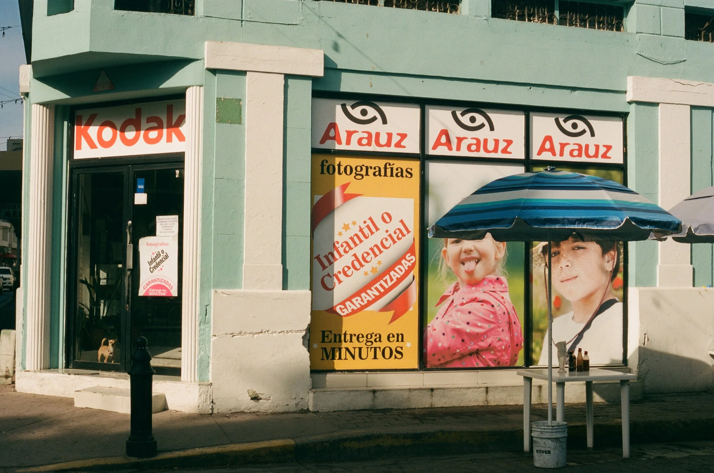 The image shows the exterior of a store with signs for Kodak and Arauz photography services. There are advertisements in Spanish for children's and maternity photography, with images of a smiling young girl and a boy. A blue striped umbrella partly c