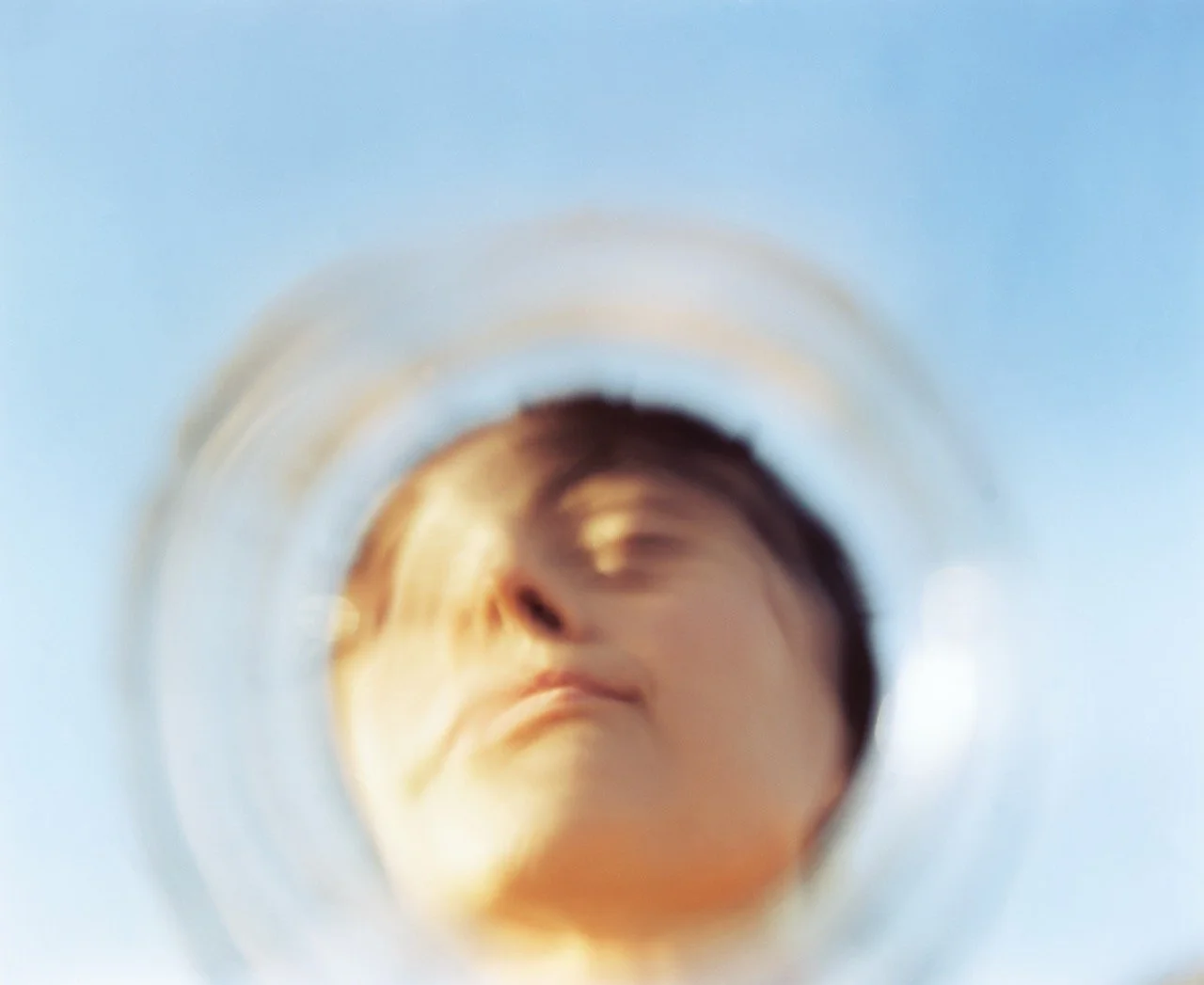 Close-up view through a glass, showing a person's face reflected, with visible lips and nose, against a light blue background.