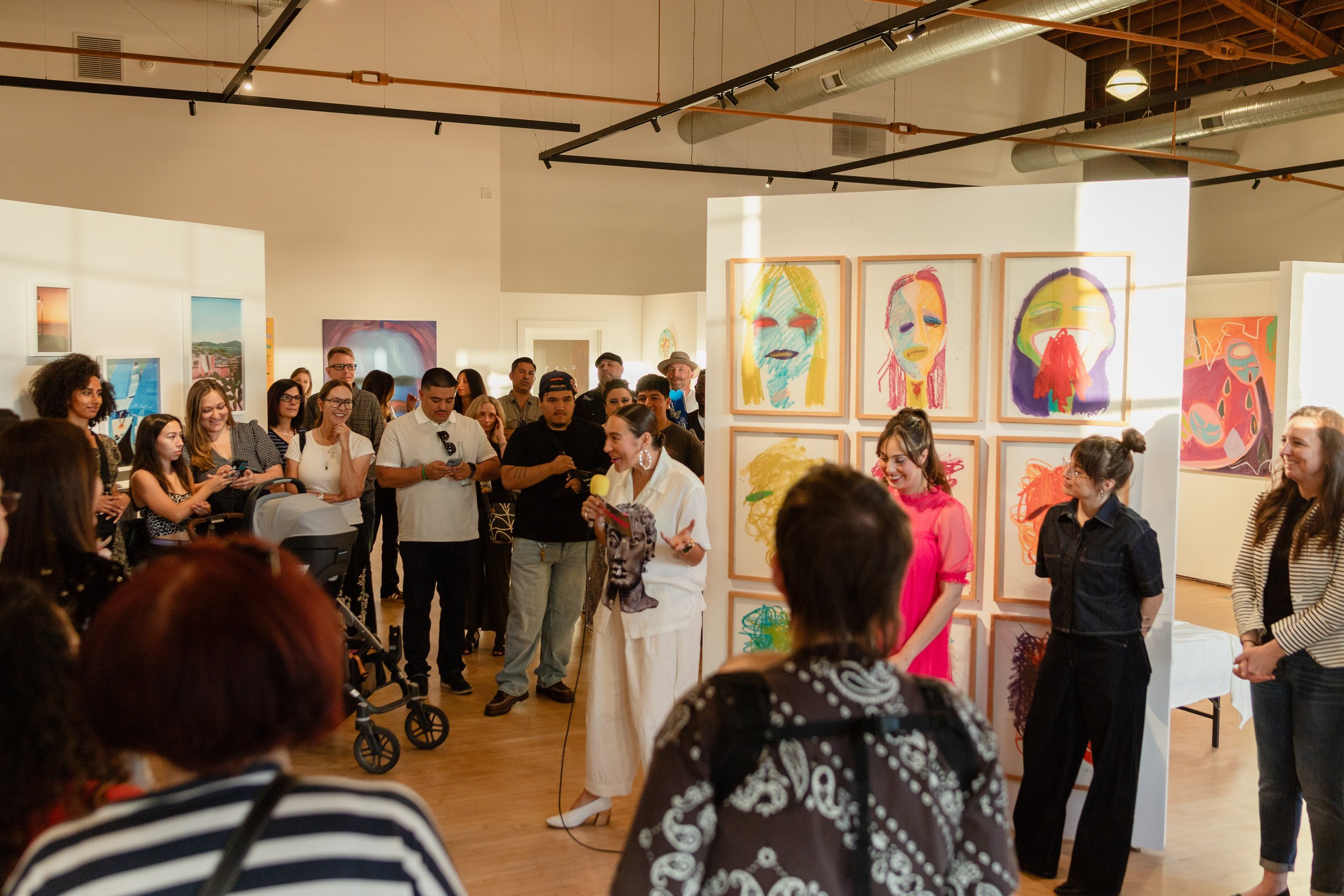 An art gallery exhibition with colorful portrait paintings on display. People are gathered, some listening to a woman speaking into a microphone, others observing the art, in a well-lit room with modern decor.