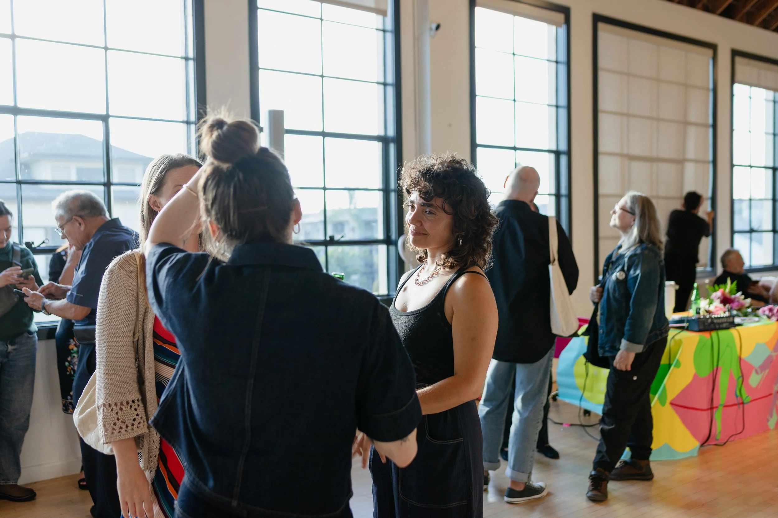 People mingling at an indoor event with large windows, colorful table displays, and vibrant decorations.