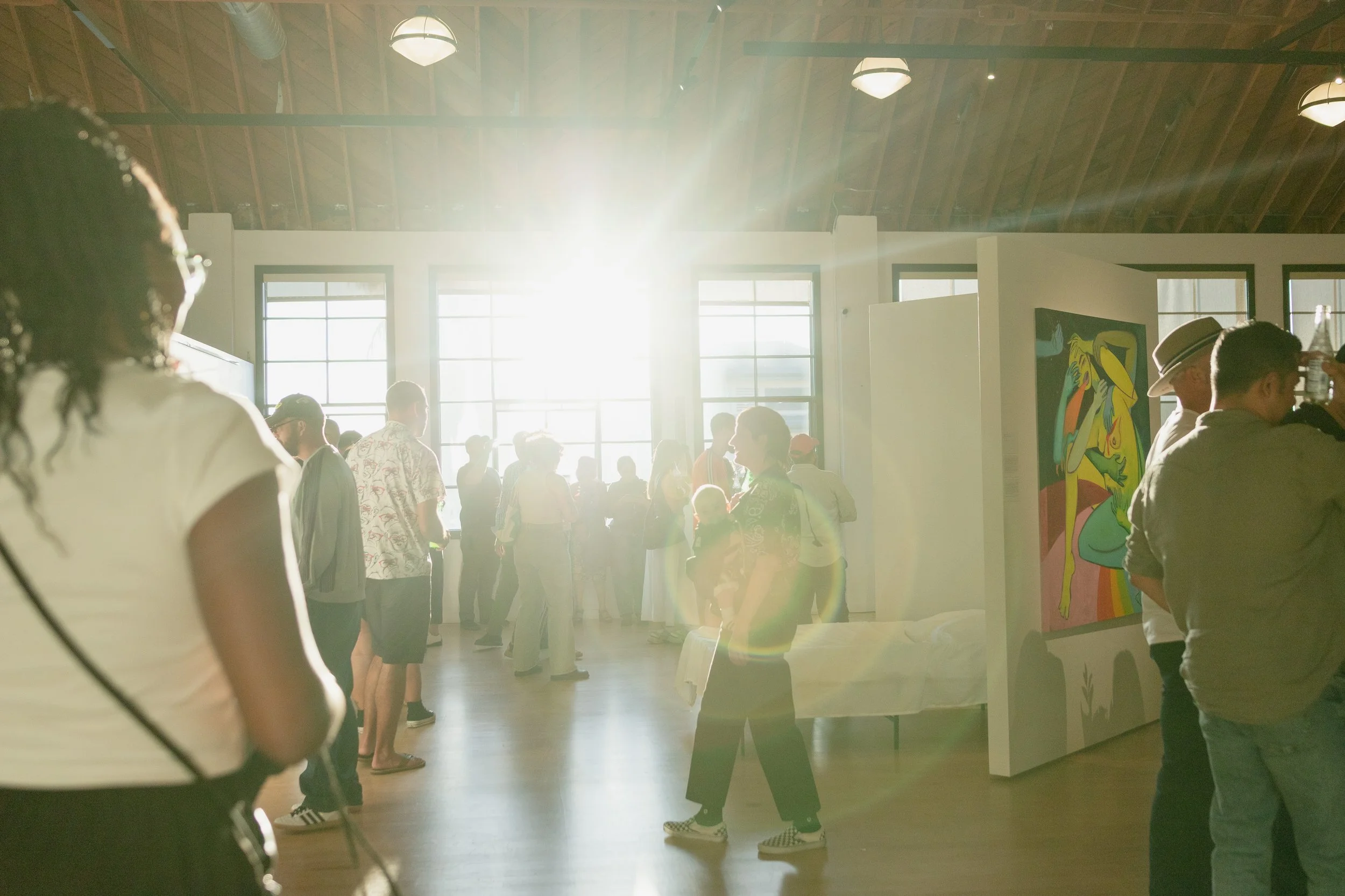 People gathered inside an art gallery with sunlight streaming through large windows, viewing colorful artwork on display.