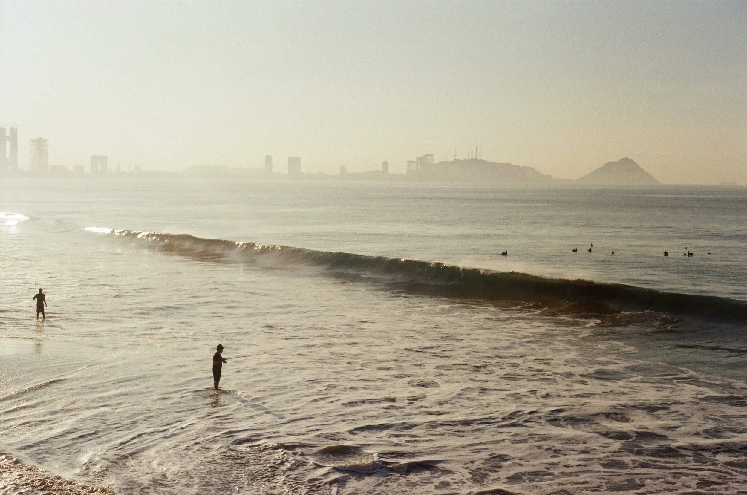 People walking and standing along the shore with waves breaking, city skyline with tall buildings, and a hill with antennas in the background