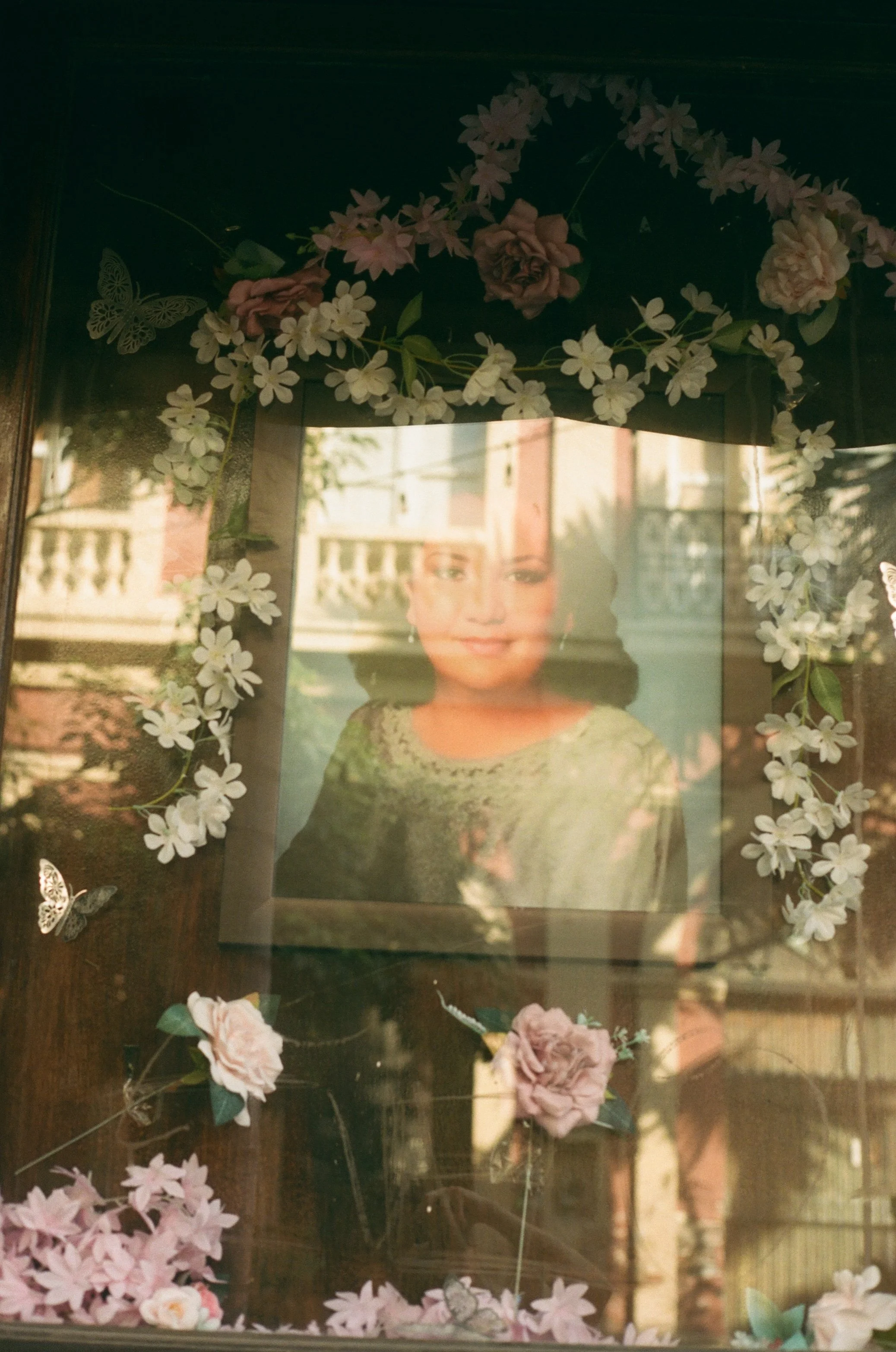 A framed portrait of a woman is surrounded by pink and white flowers and butterfly decorations on a dark surface, with reflections of the background visible in the glass.
