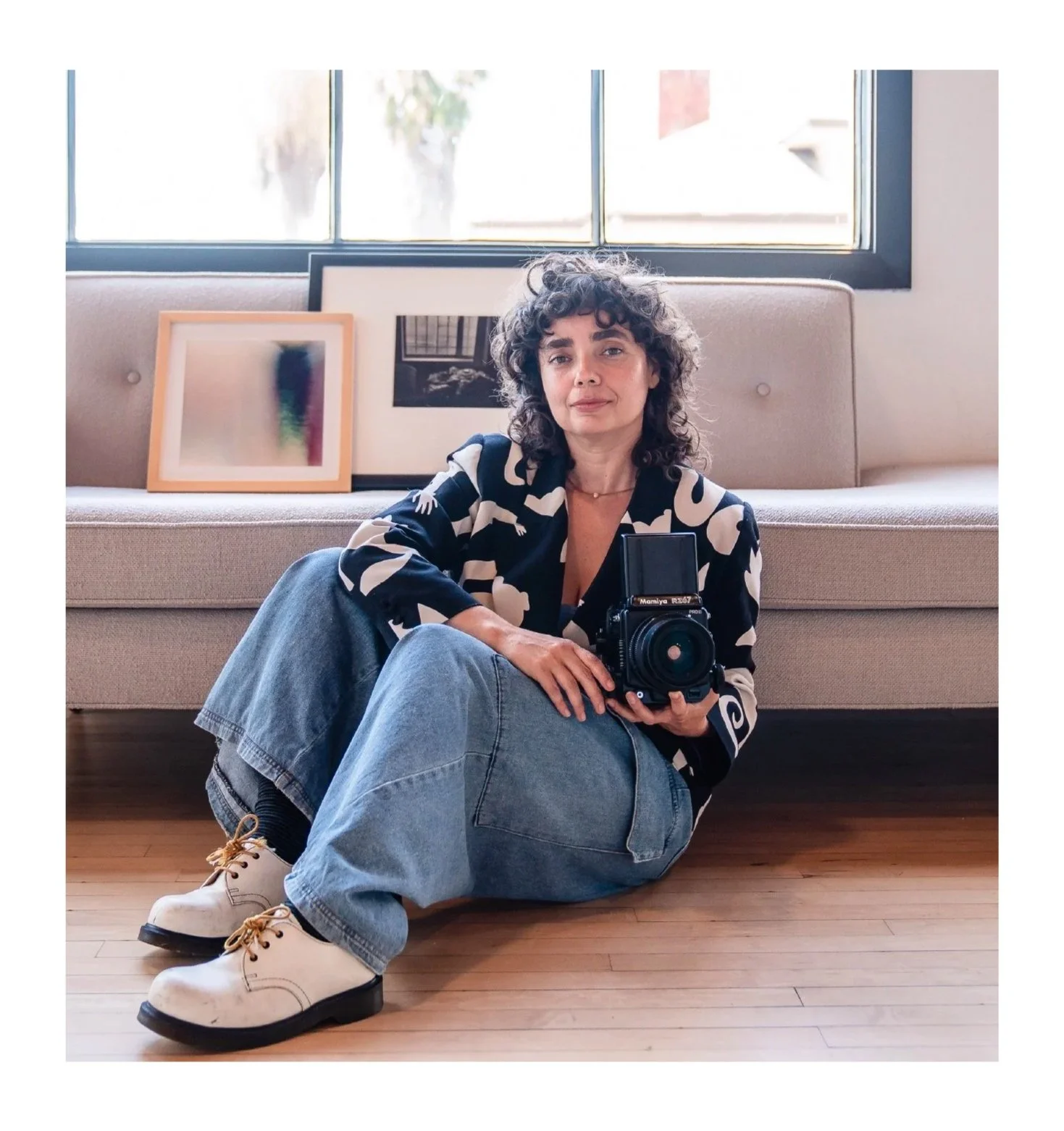 A woman with curly hair, wearing a black and white patterned shirt, light blue jeans, and white boots, sitting on a wooden floor in front of a beige couch with framed artwork behind her, holding a vintage camera.