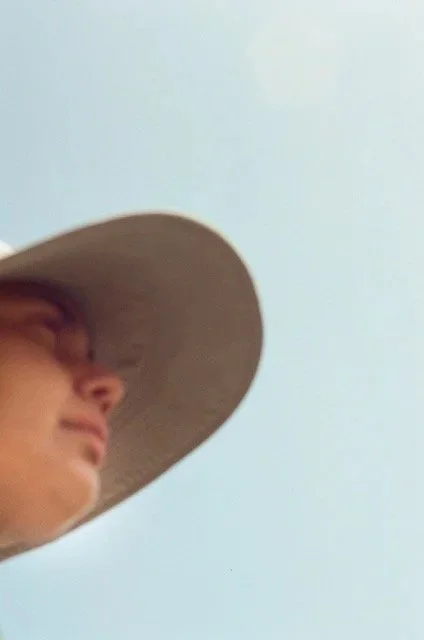 Close-up of a person wearing a wide-brimmed hat, facing slightly to the left, against a pale blue sky background.