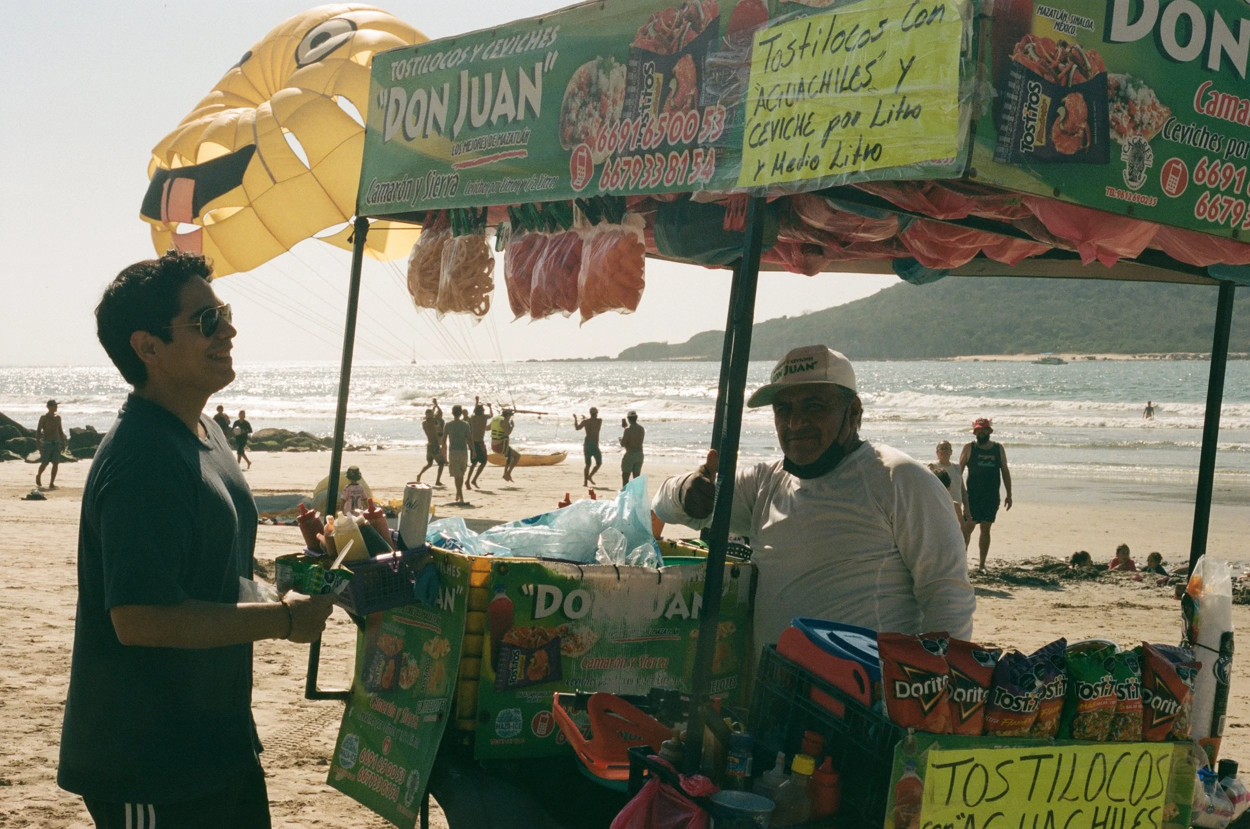 A street vendor stand on the beach selling tostadas with shrimp and avocado, with a yellow hot air balloon in the background and people walking along the shore.