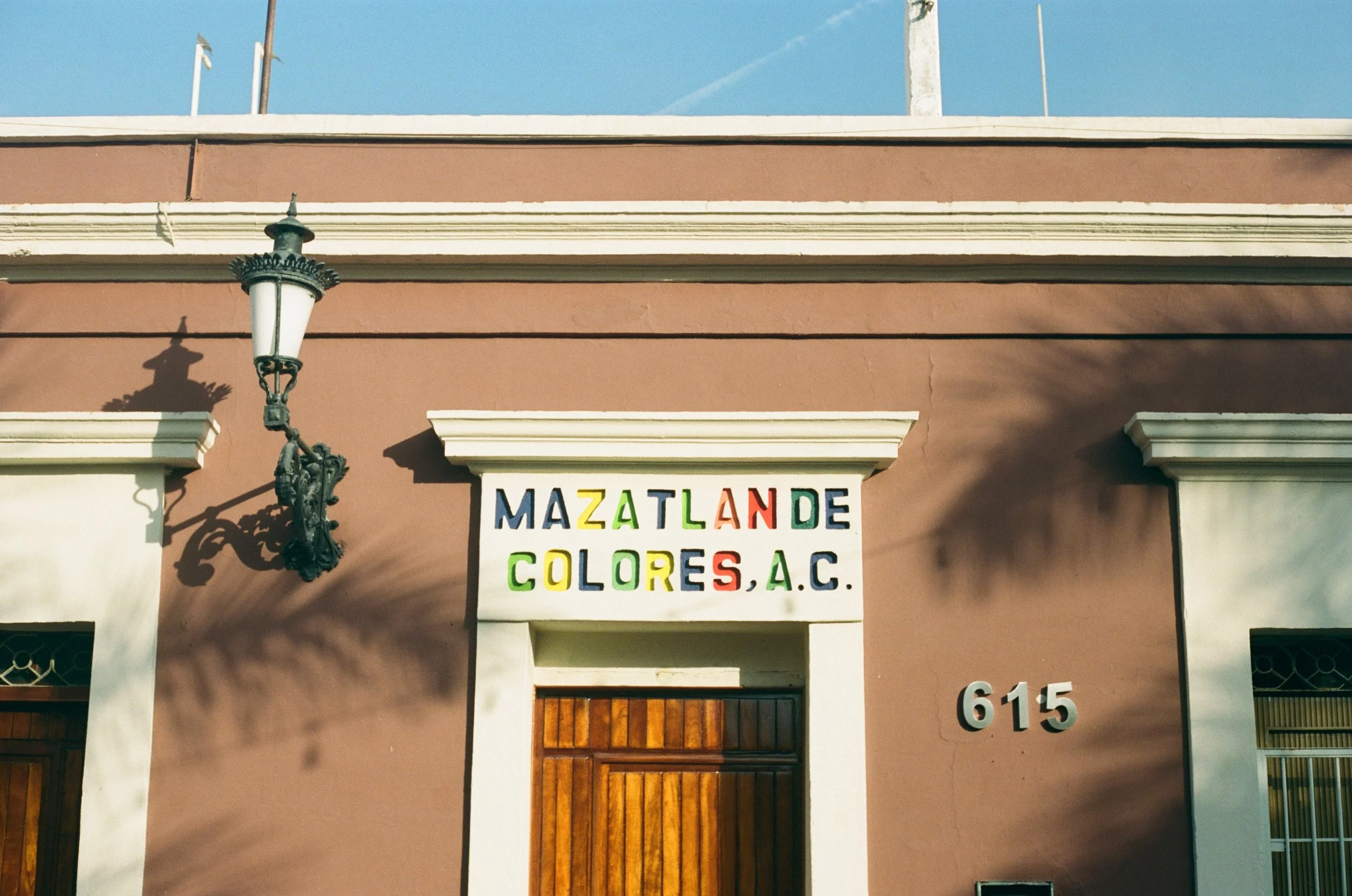 A building facade with a vintage black street lamp and a colorful sign that reads 'Mazatlán Colores A.C.' above an arched entrance. The building has a pinkish wall with white decorative accents and the number 615.