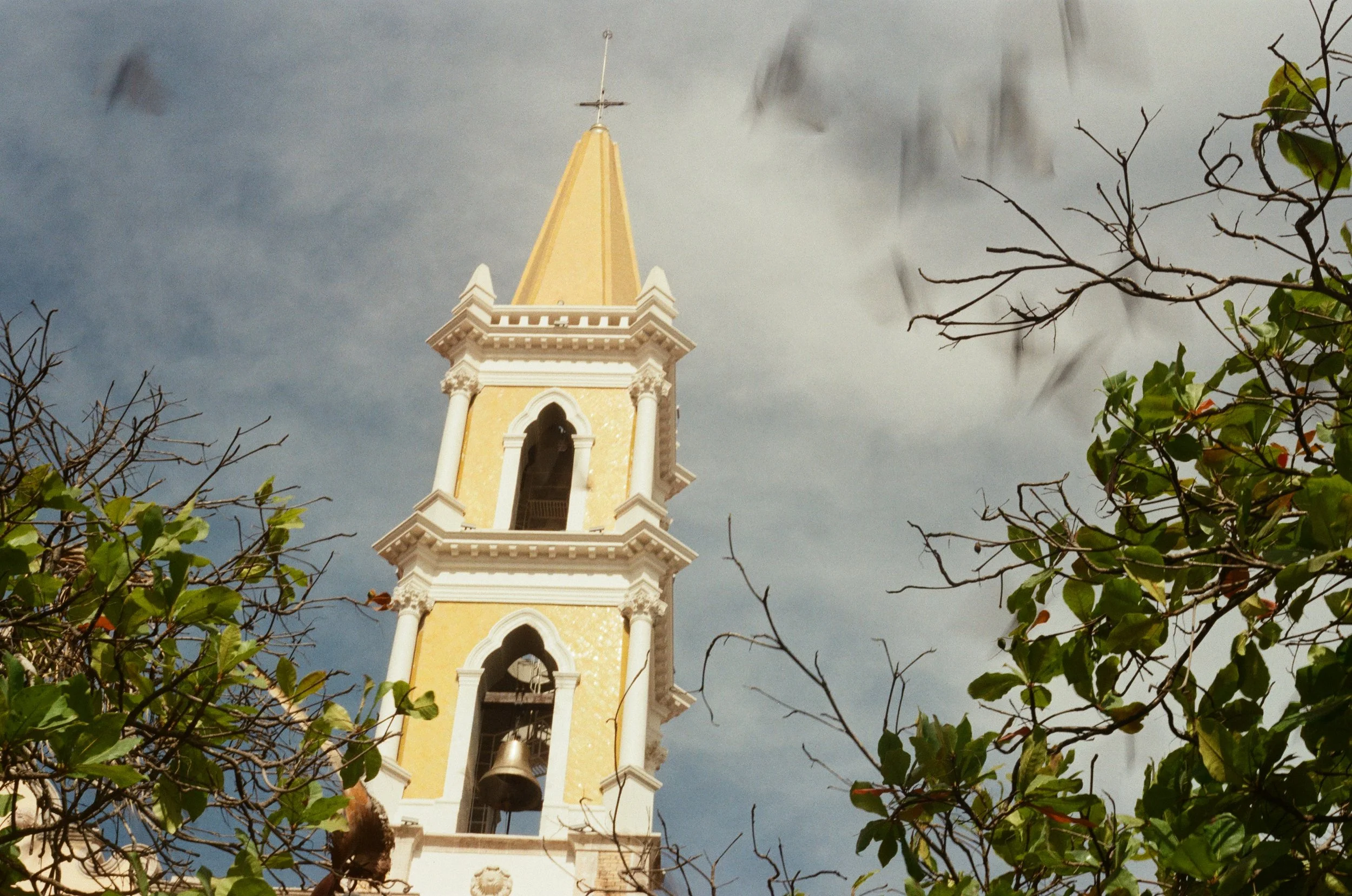 A yellow and white church steeple with a bell, viewed through tree branches against a cloudy sky.