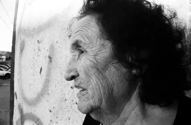 A black and white photo of an elderly woman with curly dark hair, looking to her left, standing outdoors near a wall with peeling paint.