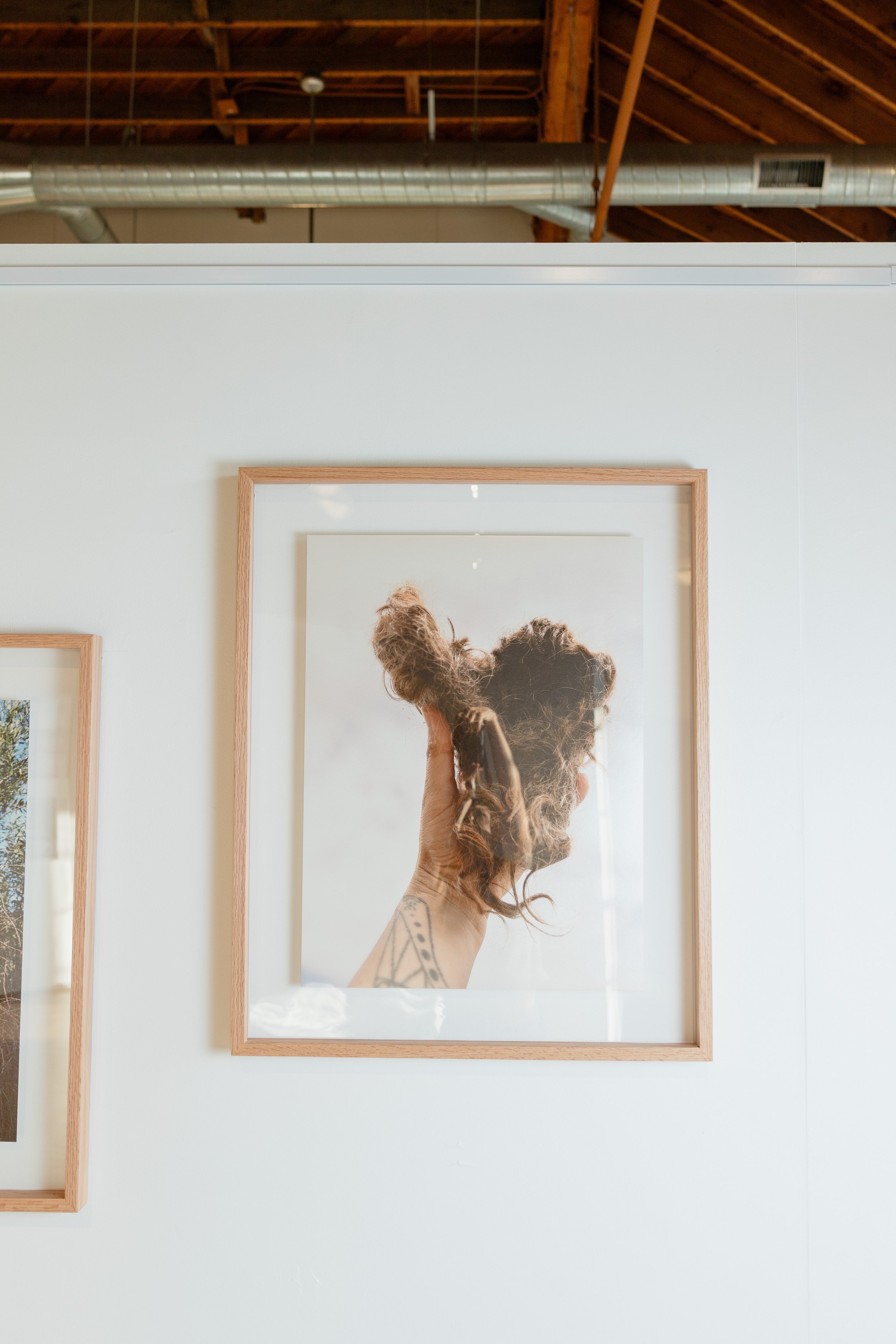 Photo of a woman with curly hair and a tattoo on her arm looking at herself in a framed mirror. The mirror is mounted on a white wall, with a large photo or artwork reflected in the background. The scene appears to be in a modern, industrial-style sp