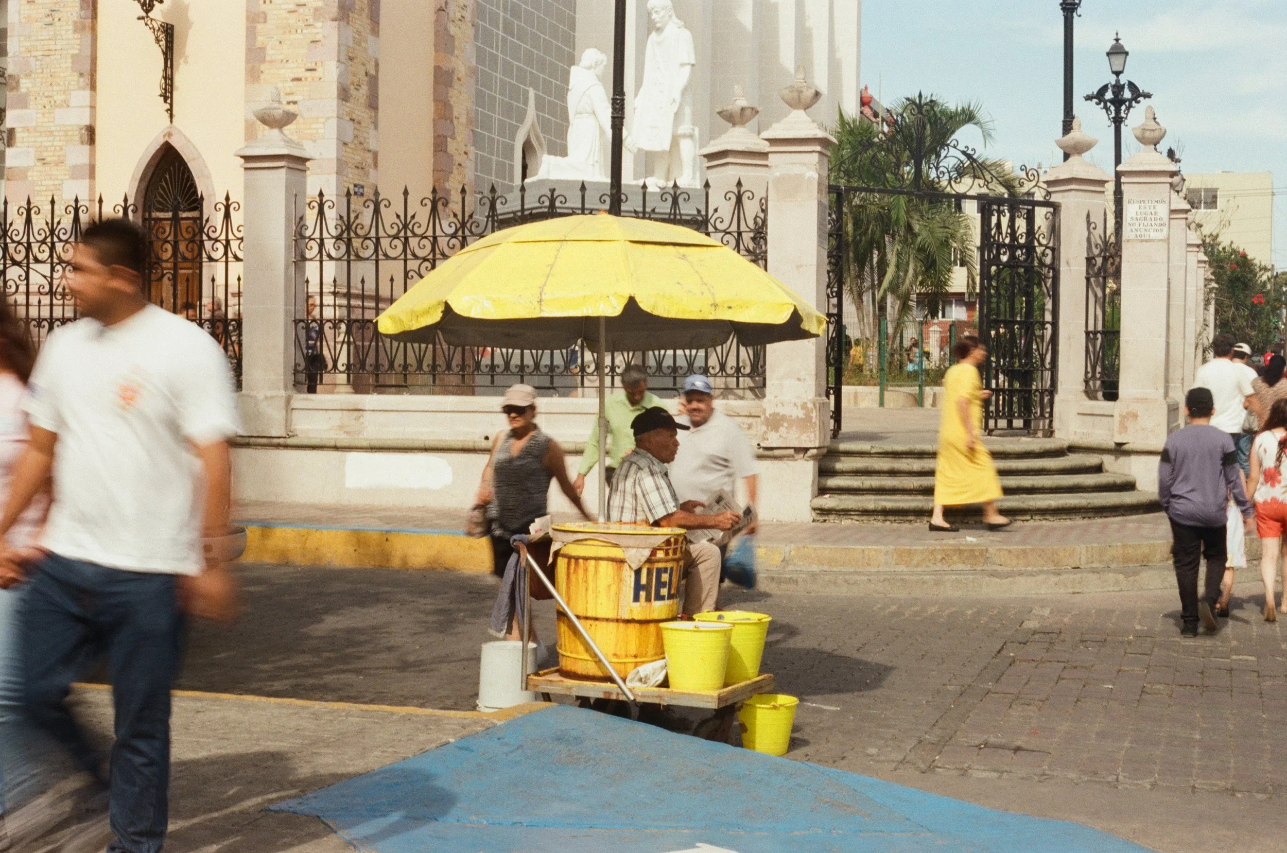 People walking and sitting in front of a church with a black wrought iron gate. A yellow umbrella shades a street vendor cart, and some people are dressed casually. There are trees and buildings in the background.