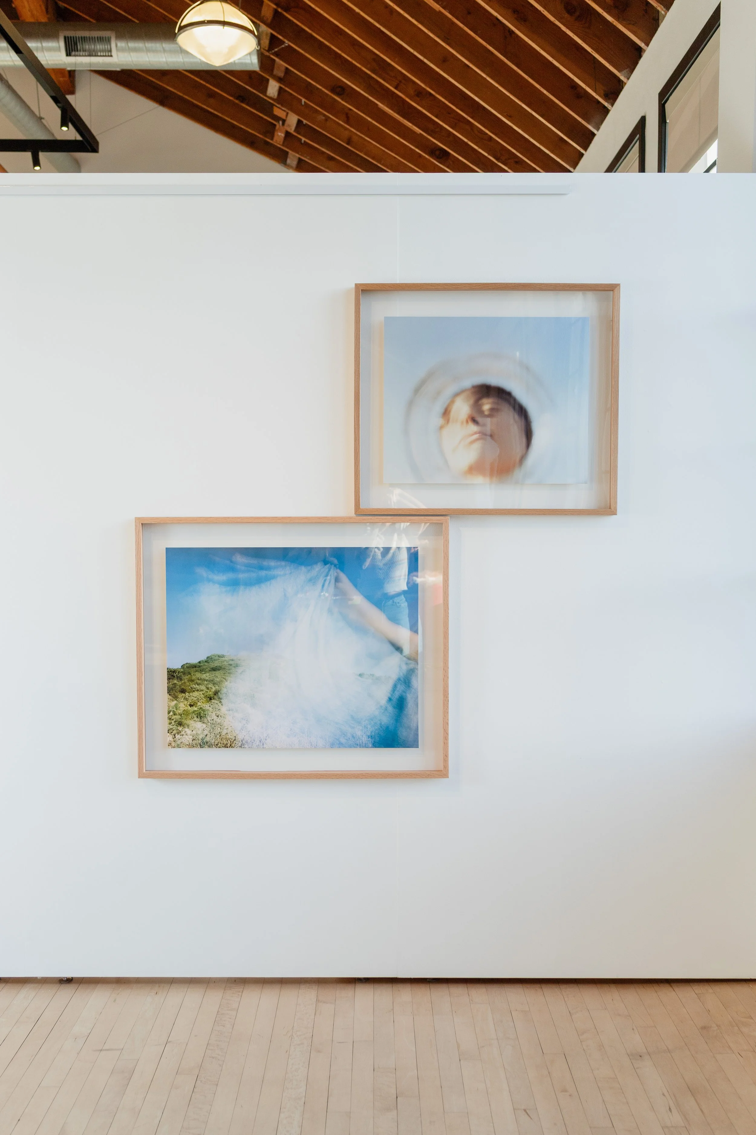 Two framed photographs hanging on a white gallery wall, with wooden floors and a wooden ceiling visible at the top, in an art gallery.