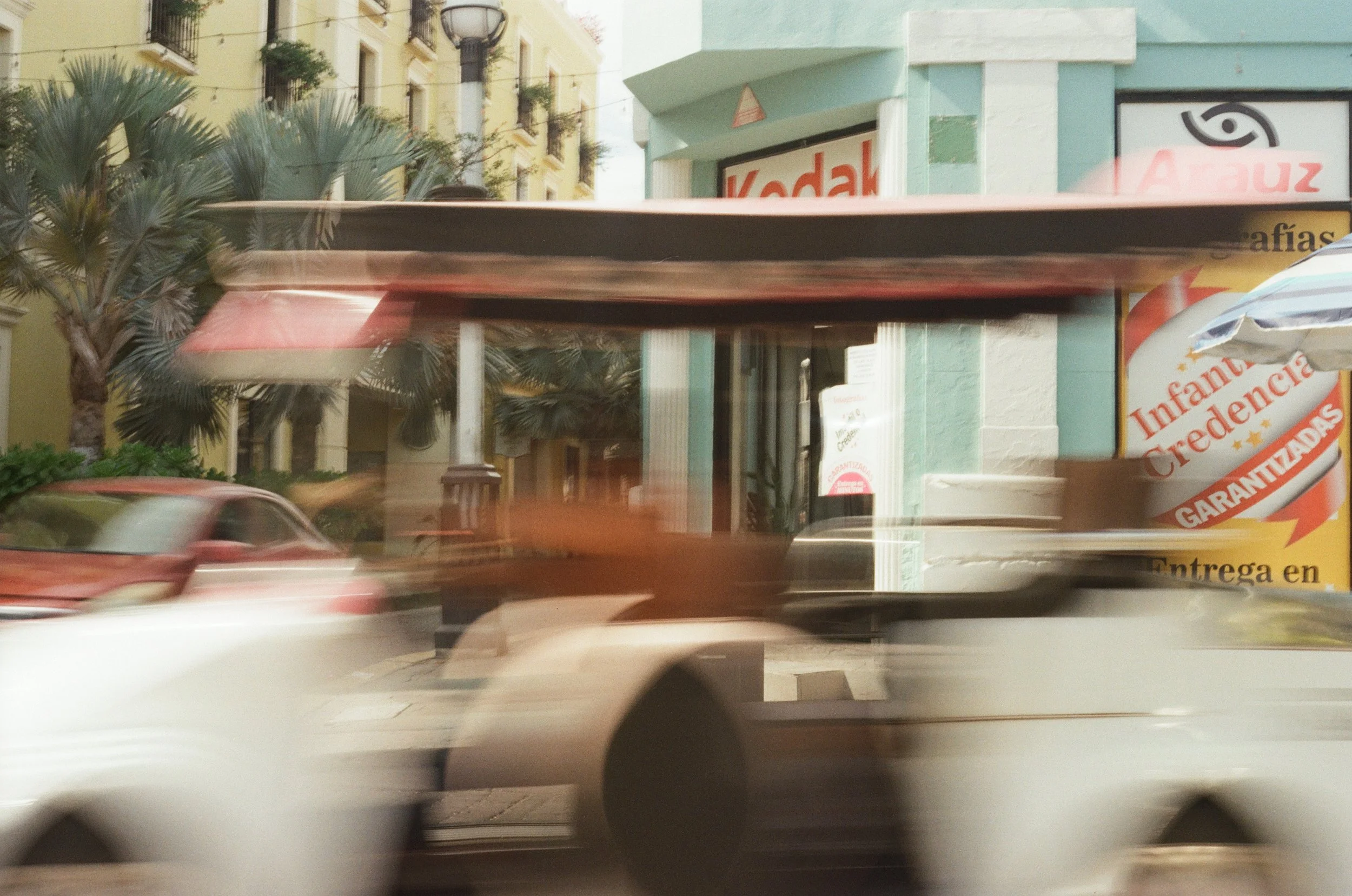 Blurred city street scene with moving vehicles, storefronts including Kodak sign, surrounding buildings, and palm trees.
