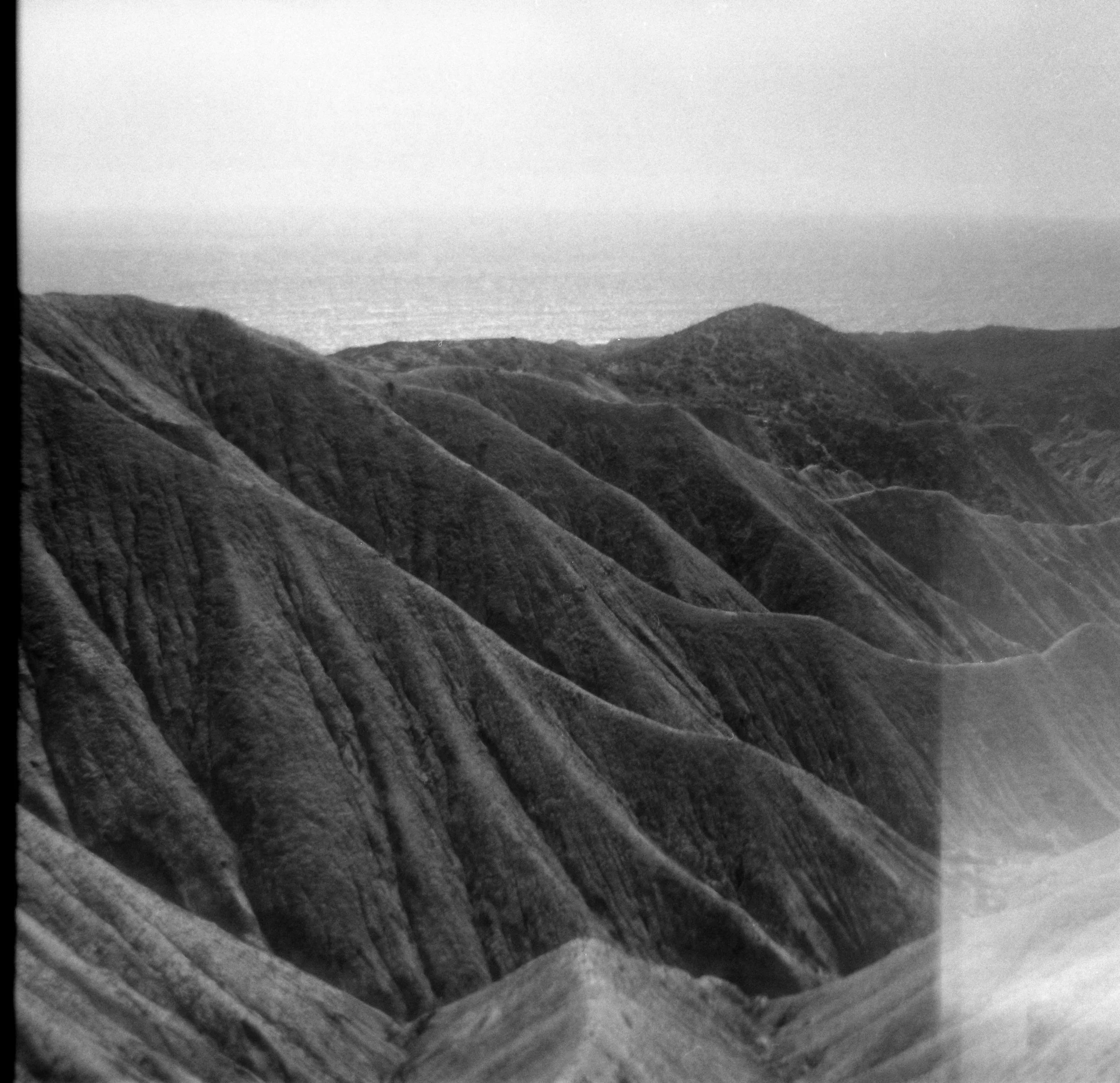 Black and white photo of mountain ridges and a body of water in the distance under a cloudy sky.