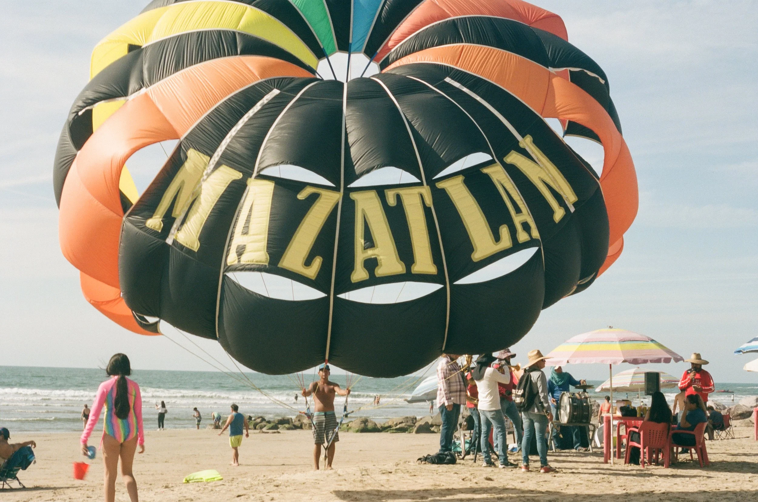 People on the beach preparing for a hot air balloon ride; the balloon is dark with yellow lettering, surrounded by umbrellas and beachgoers.