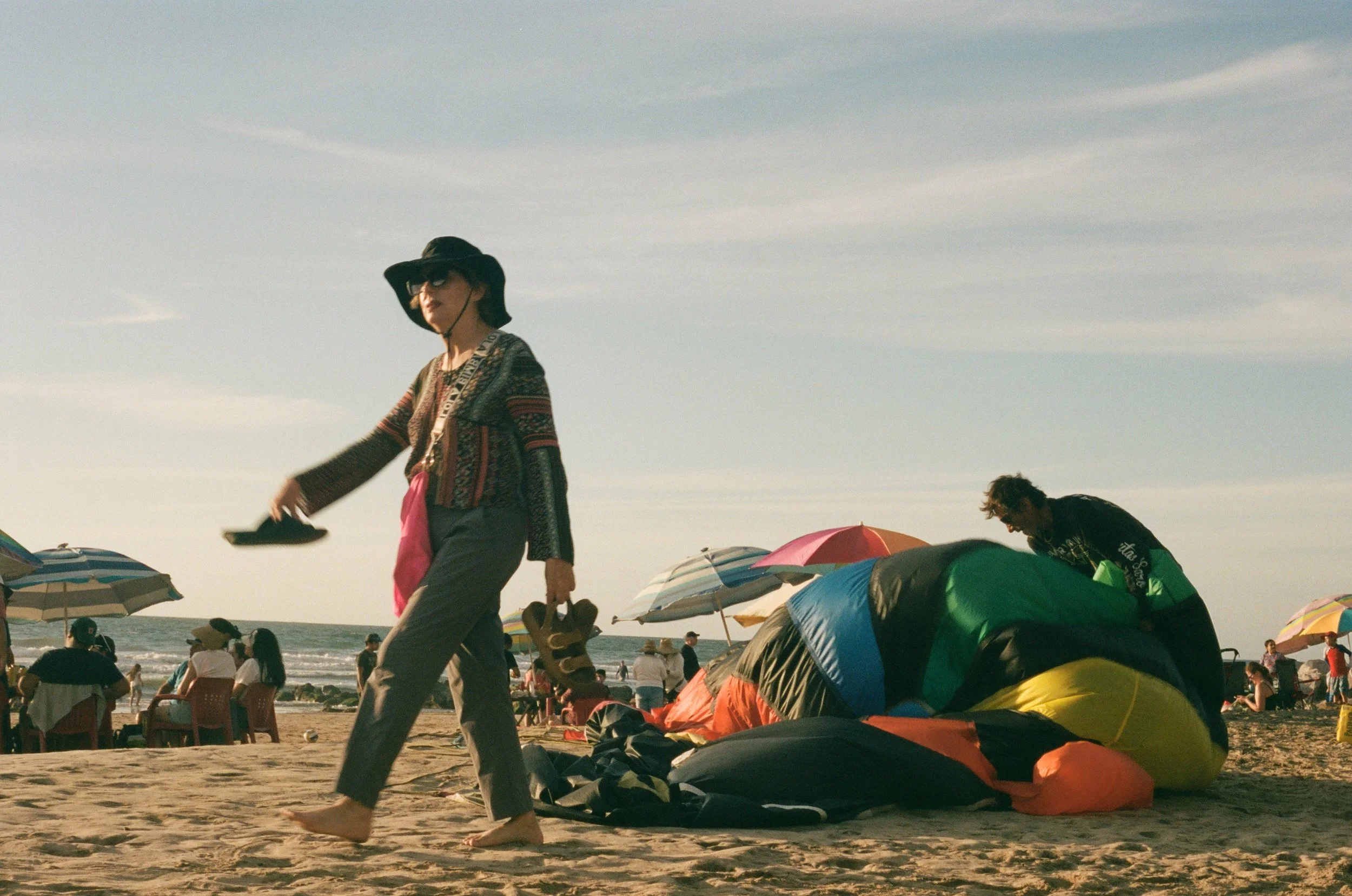 People on a crowded beach with umbrellas, a woman in a wide-brimmed hat walking, and a person packing a tent.