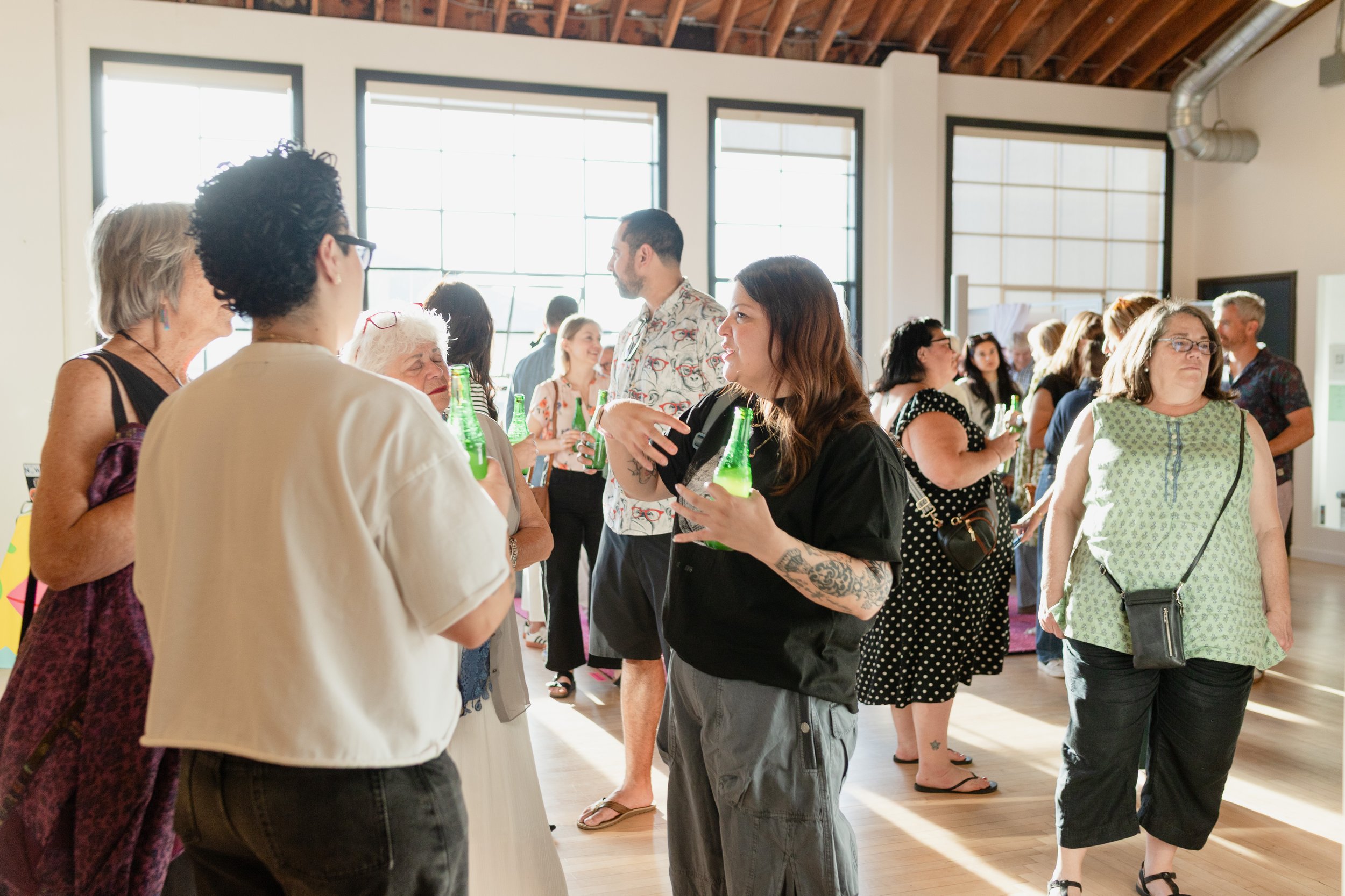 People socializing and talking at an indoor event with large windows and wooden ceiling, some holding green bottles.