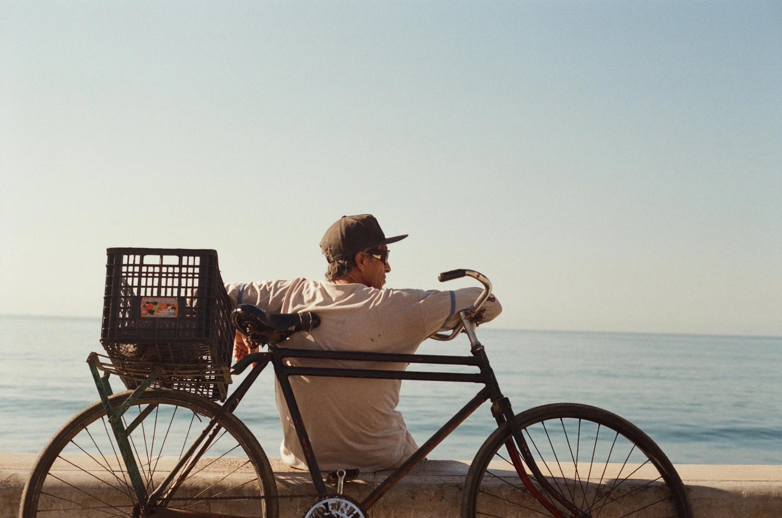 A man wearing a hat and sunglasses riding a bicycle along a beachfront path. The bicycle has a large basket at the back. The background shows the ocean and a clear sky.