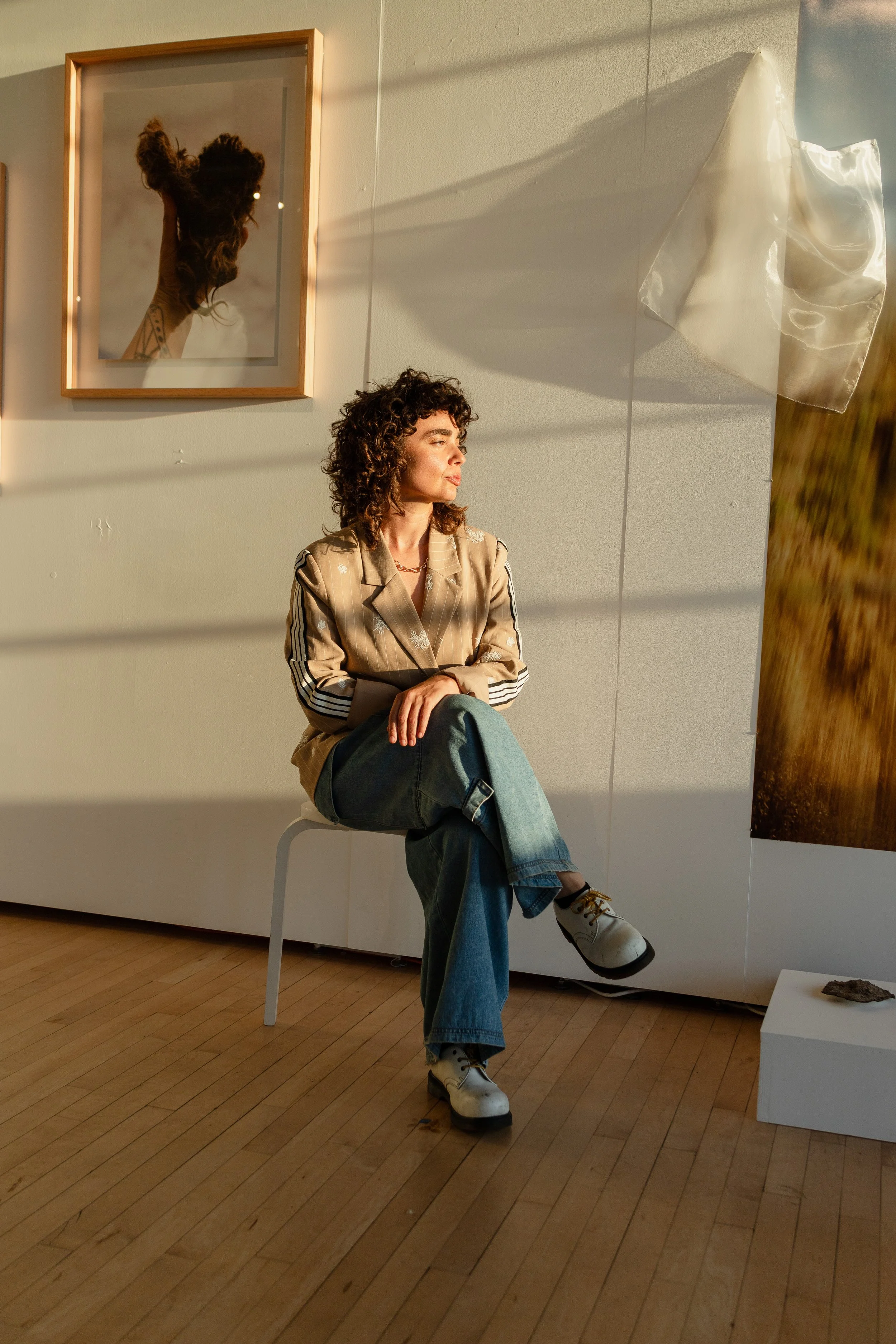 A woman with curly hair sitting on a chair in an art gallery, wearing a beige jacket with black and white striped sleeves, blue jeans, and white shoes. She is looking to her left. Behind her, there are several framed photographs on the wall, includin