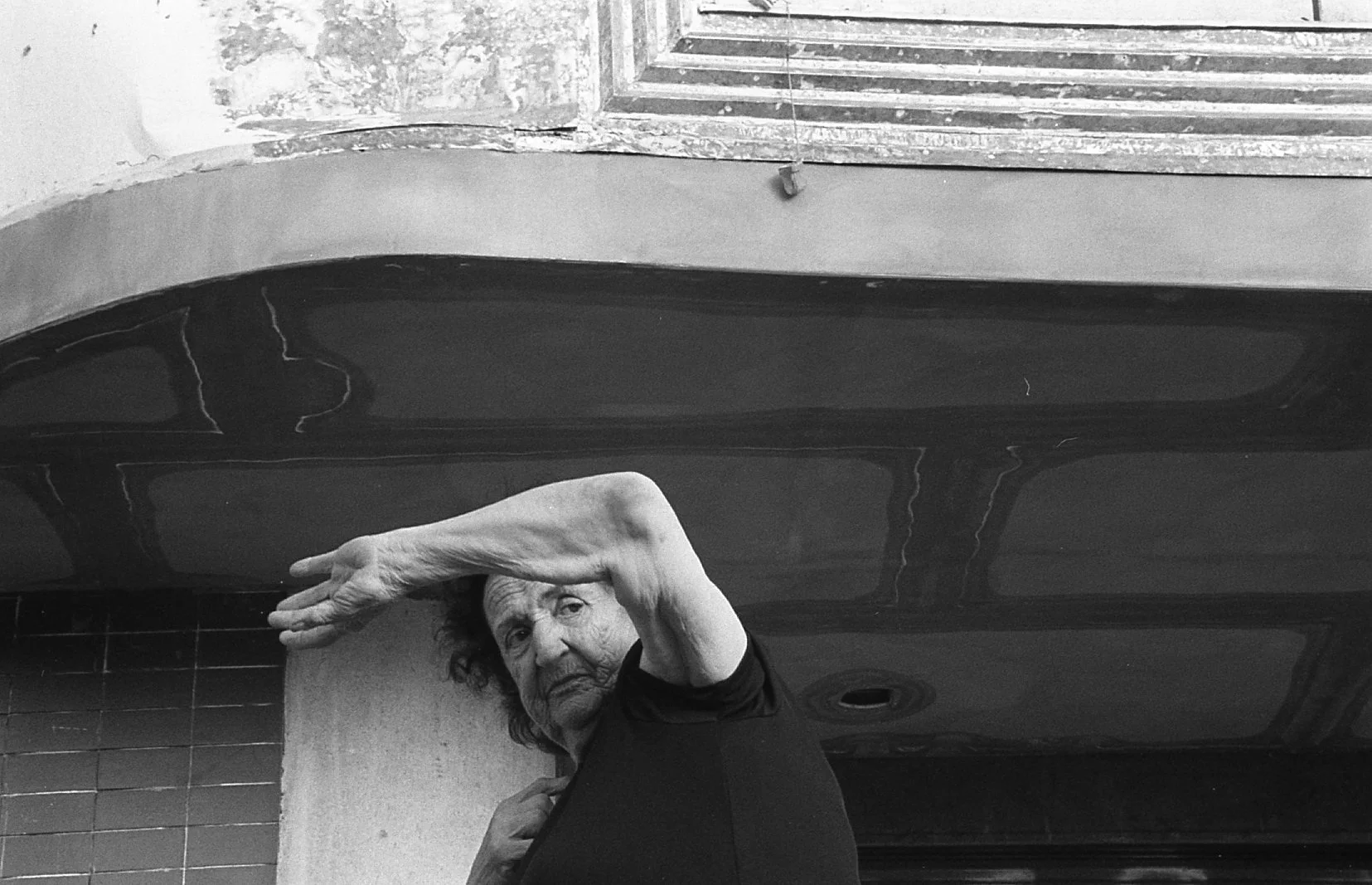 An older woman with curly hair and a black shirt is reaching into her hair with her right hand near her forehead, looking at the camera.
