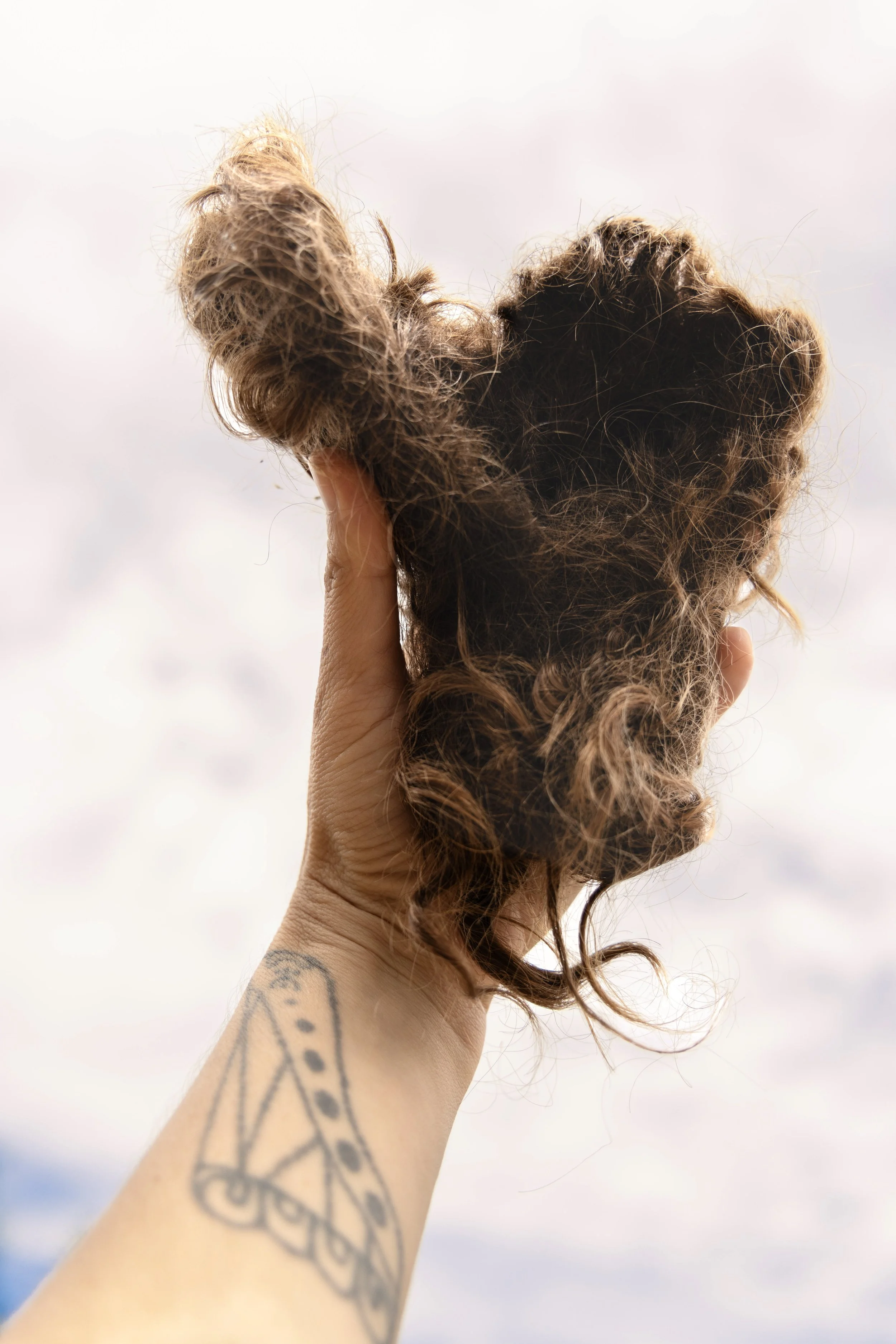 Person with tattooed arm holding a messy bun hairstyle outdoors against cloudy sky.
