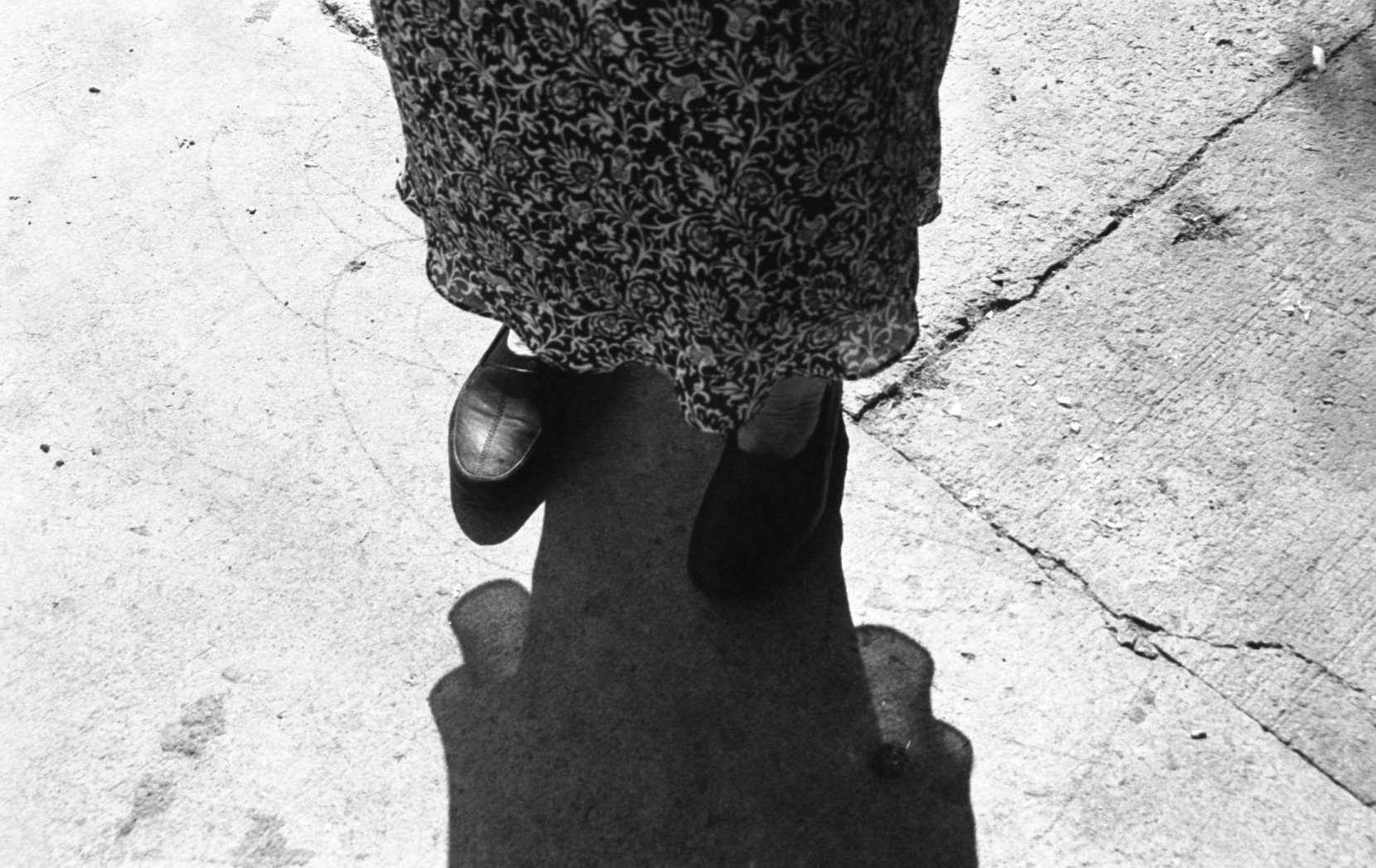 Black and white photo of a person standing on a sidewalk, wearing a floral dress and black shoes, with their own shadow cast on the ground.