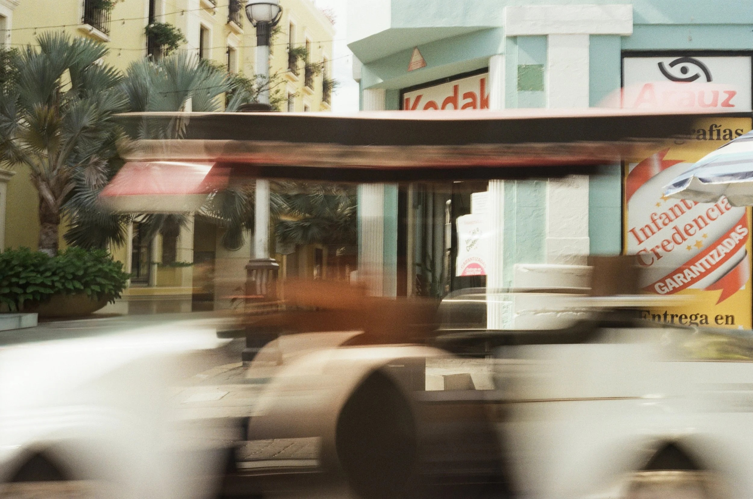 A blurred city scene with a building that has a yellow facade and balconies, reflecting some greenery. There is a palm tree and some bushes in the foreground. Part of a storefront with a sign that says "Kodal" and a large advertisement for children's medicine or vitamins, featuring bright colors and text in Spanish.