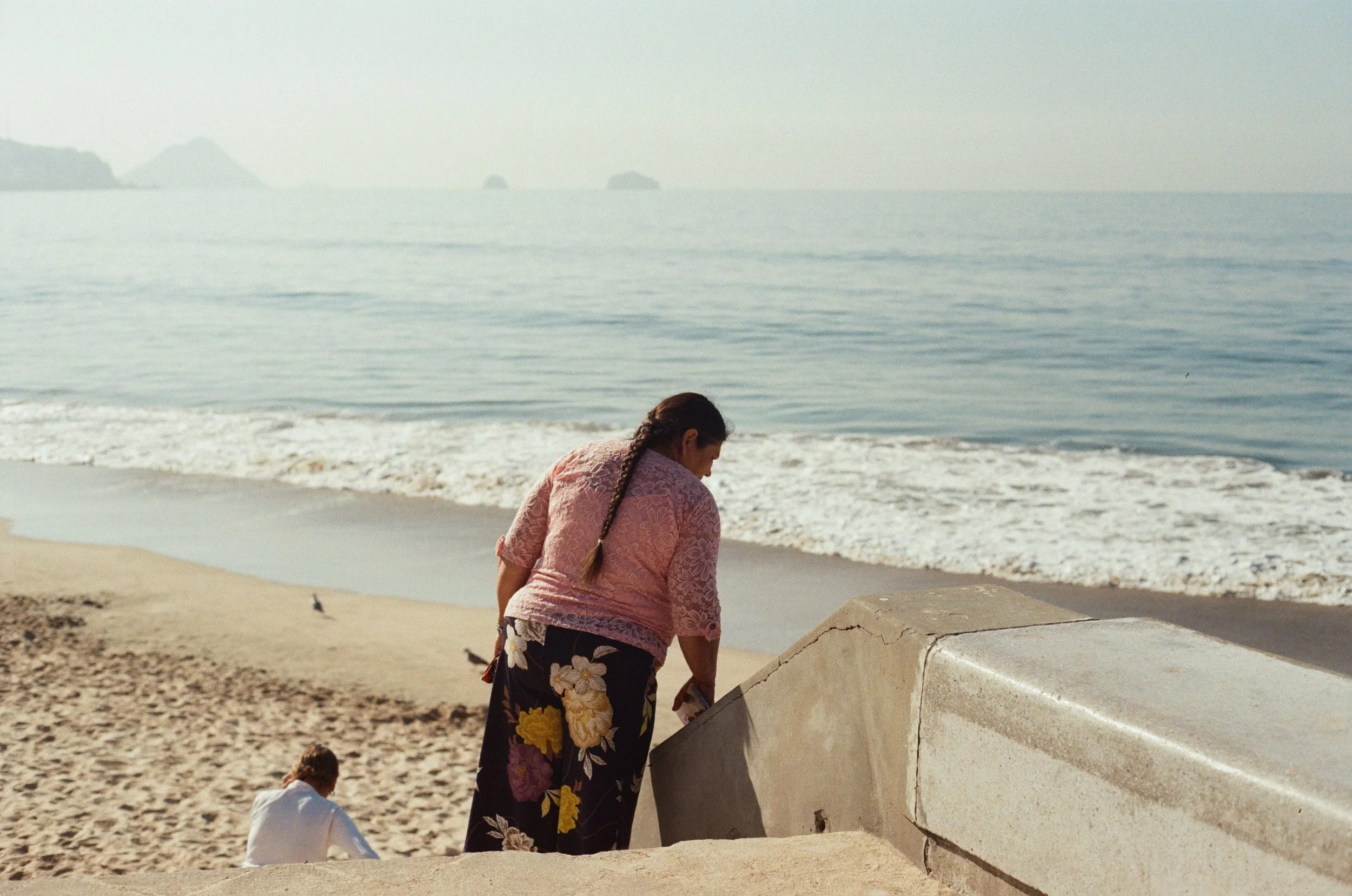 A woman with long hair in a braid wearing a pink lace top and floral skirt, leaning over a concrete structure on a beach, with two children playing nearby and the ocean in the background.