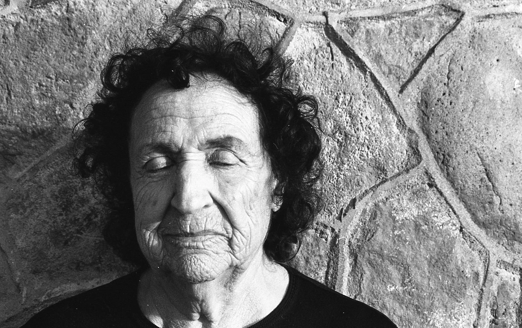 A black and white photo of an elderly woman with curled hair and closed eyes, standing against a stone wall with a star engraved in it.