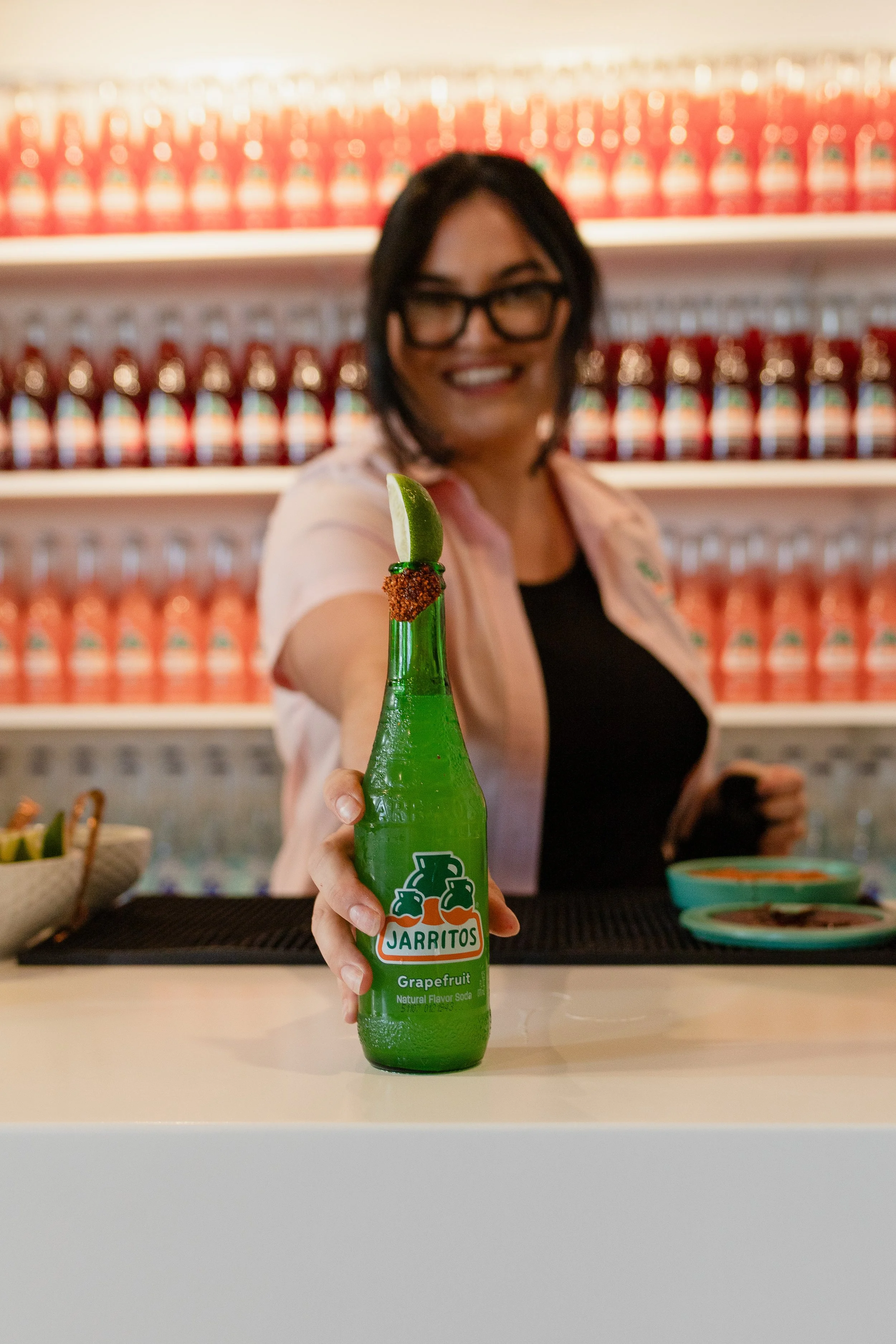 A woman with glasses smiling and holding a bottle of Jarritos grapefruit soda in front of a backdrop of stacked bottles on shelves.