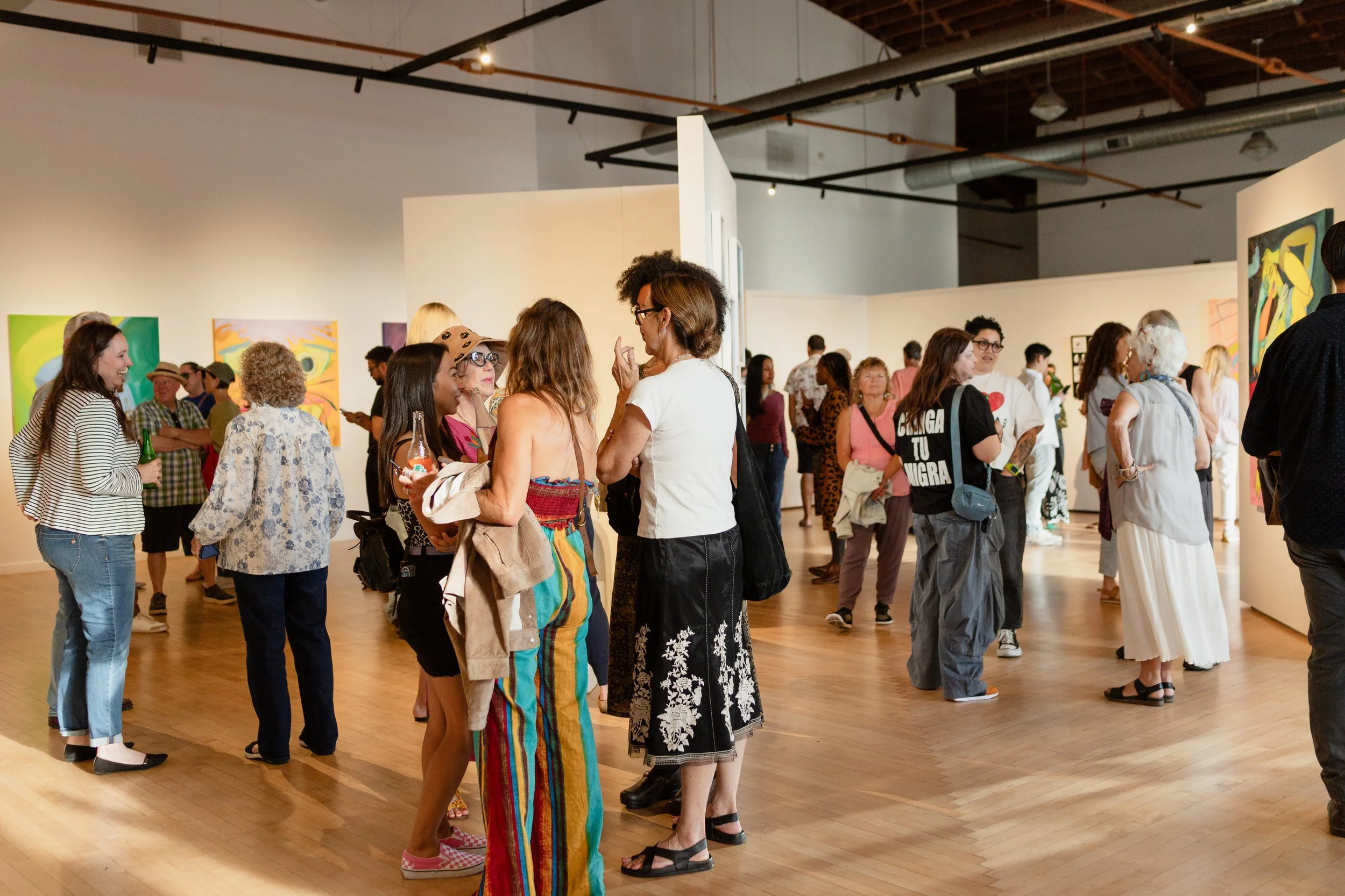 People gathered in an art gallery, engaging in conversation and viewing colorful abstract paintings on the walls.