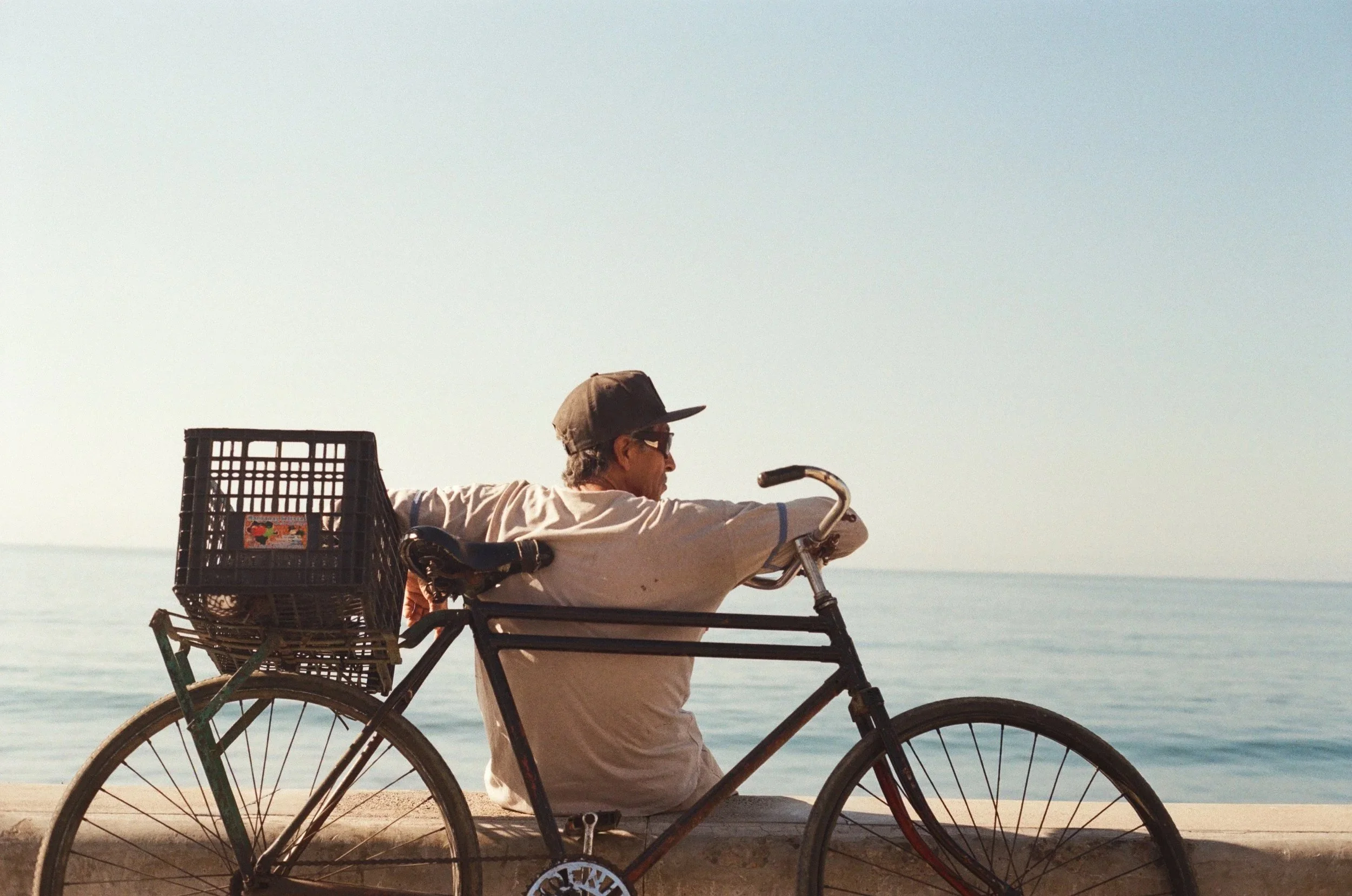 A man riding a bicycle along the beach with the ocean in the background, wearing a cap and glasses, and pulling a large crate on the back of the bike.