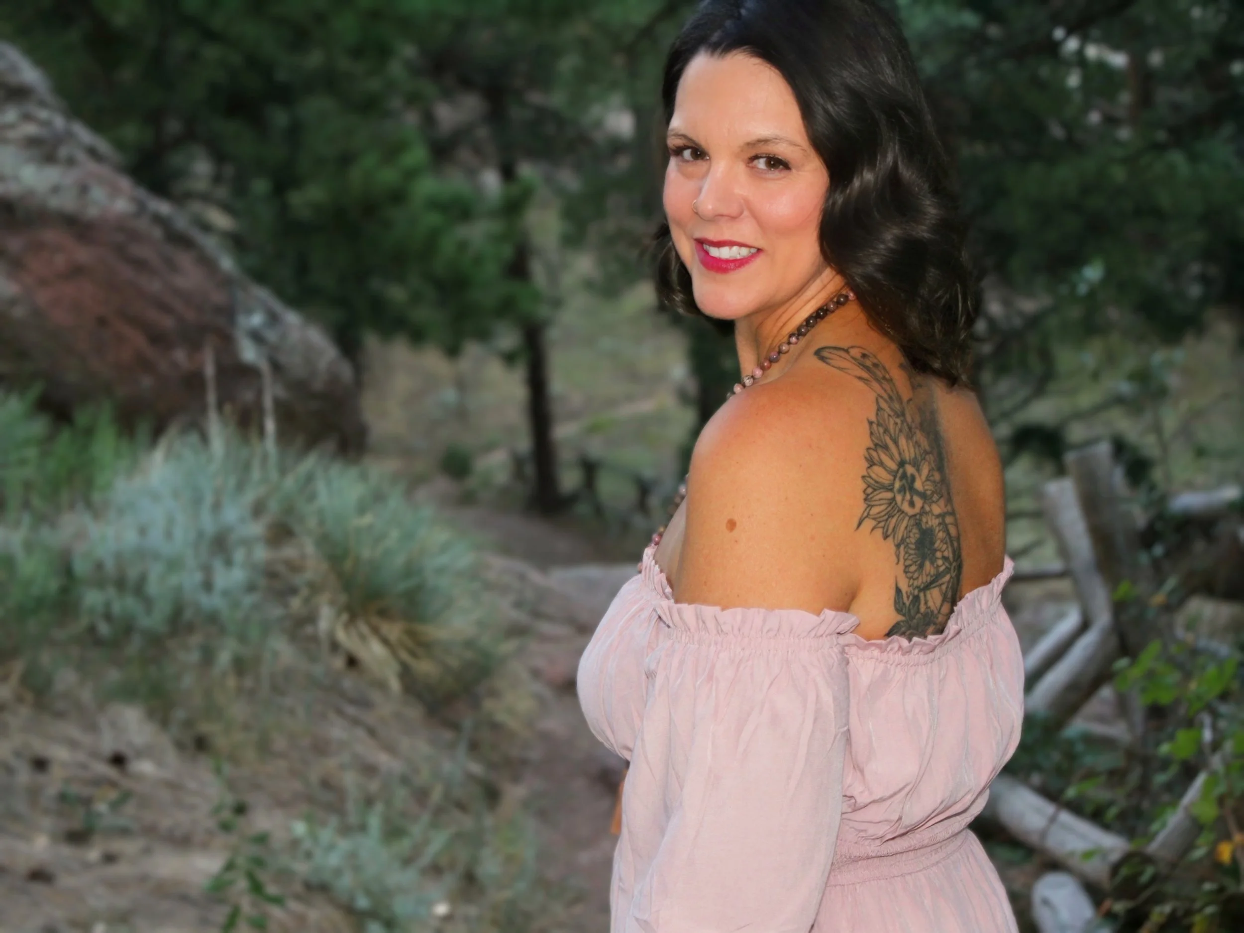A woman with black hair and a tattoo on her shoulder smiling outdoors with a blurred green background.