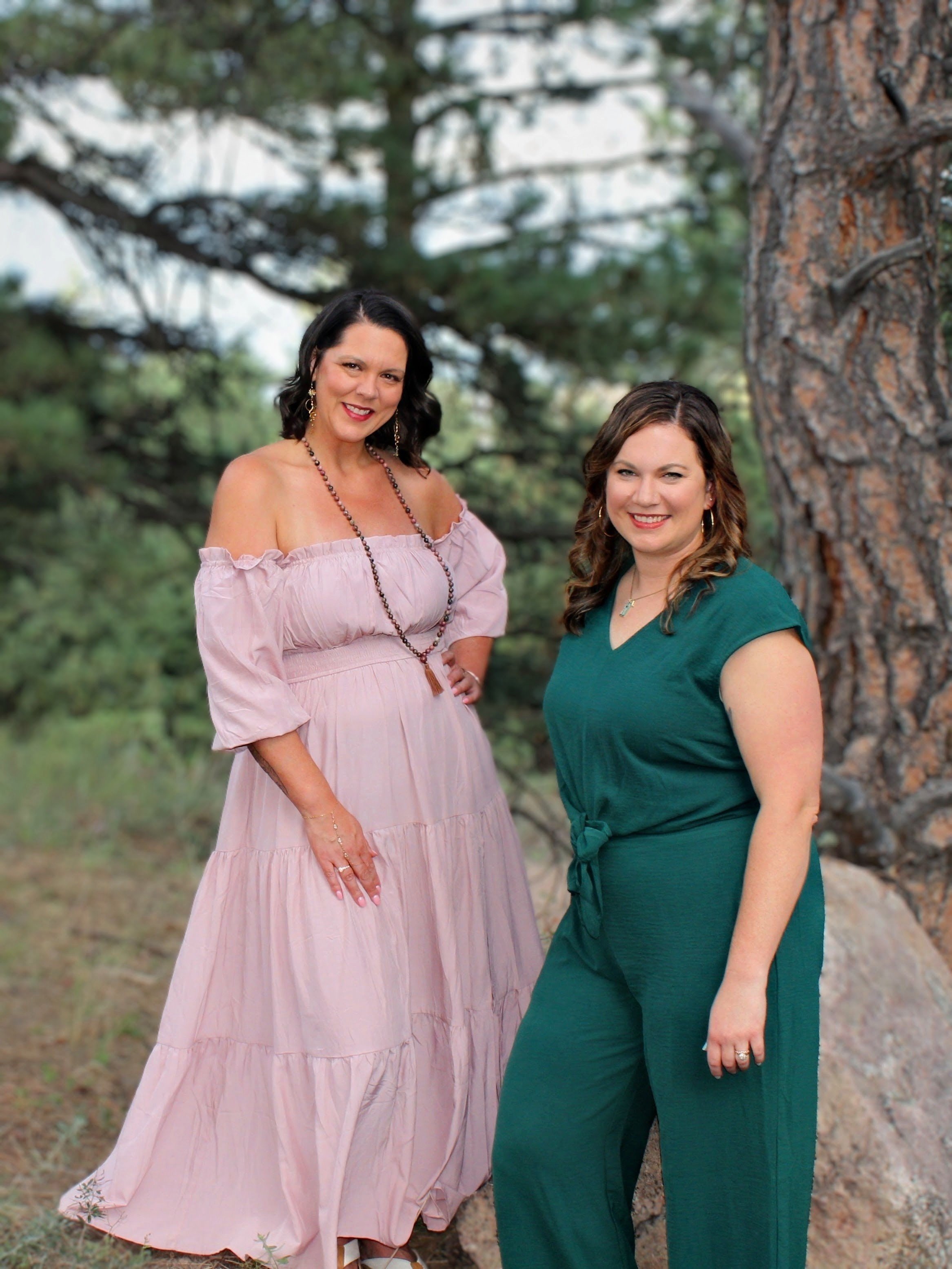 Two women standing outdoors next to a tree, smiling at the camera, with green foliage and tree branches in the background.
