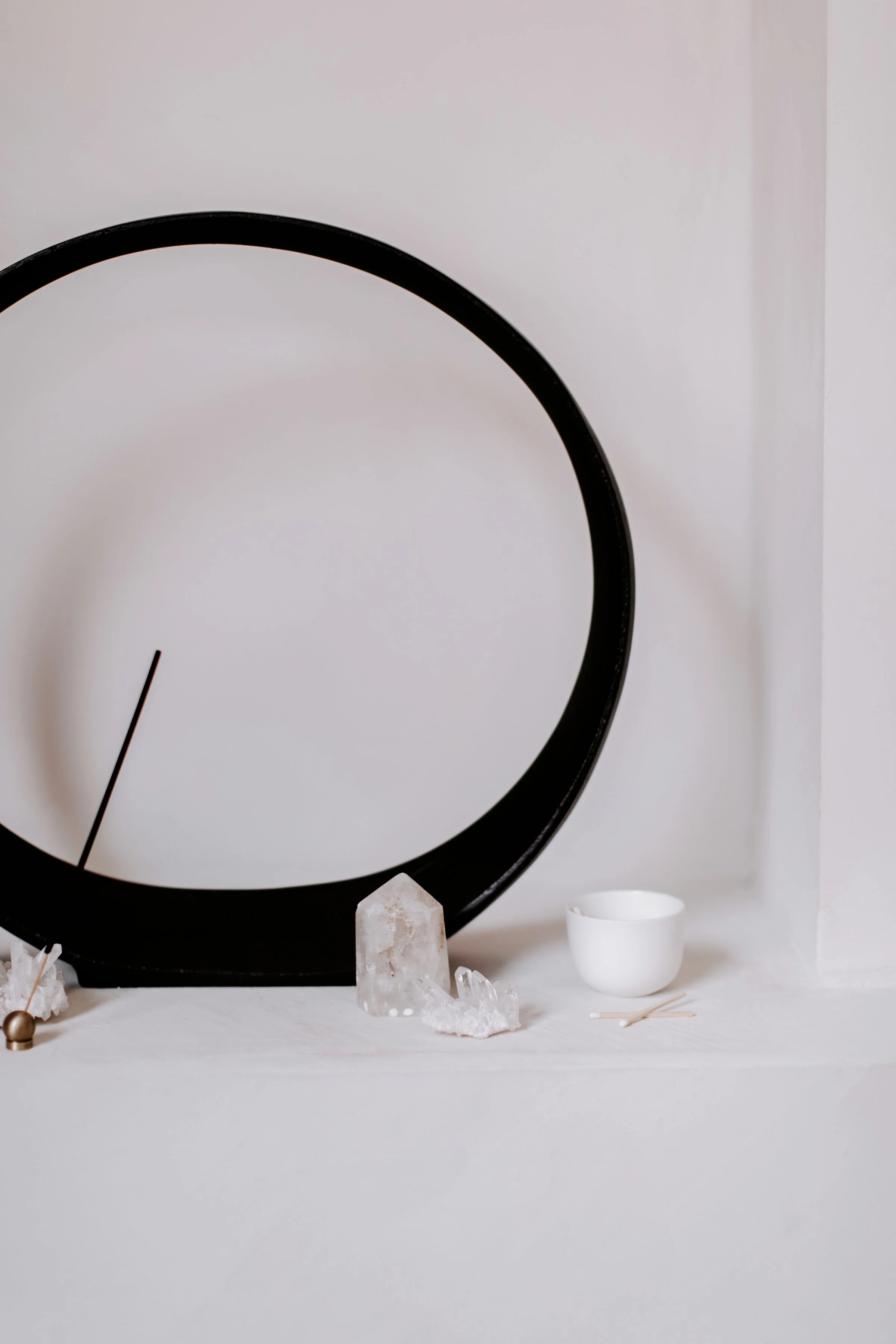 Minimalist still life arrangement with a round black clock frame, clear quartz crystals, a white bowl, and a small white cup, set on a white surface against a light-colored background.