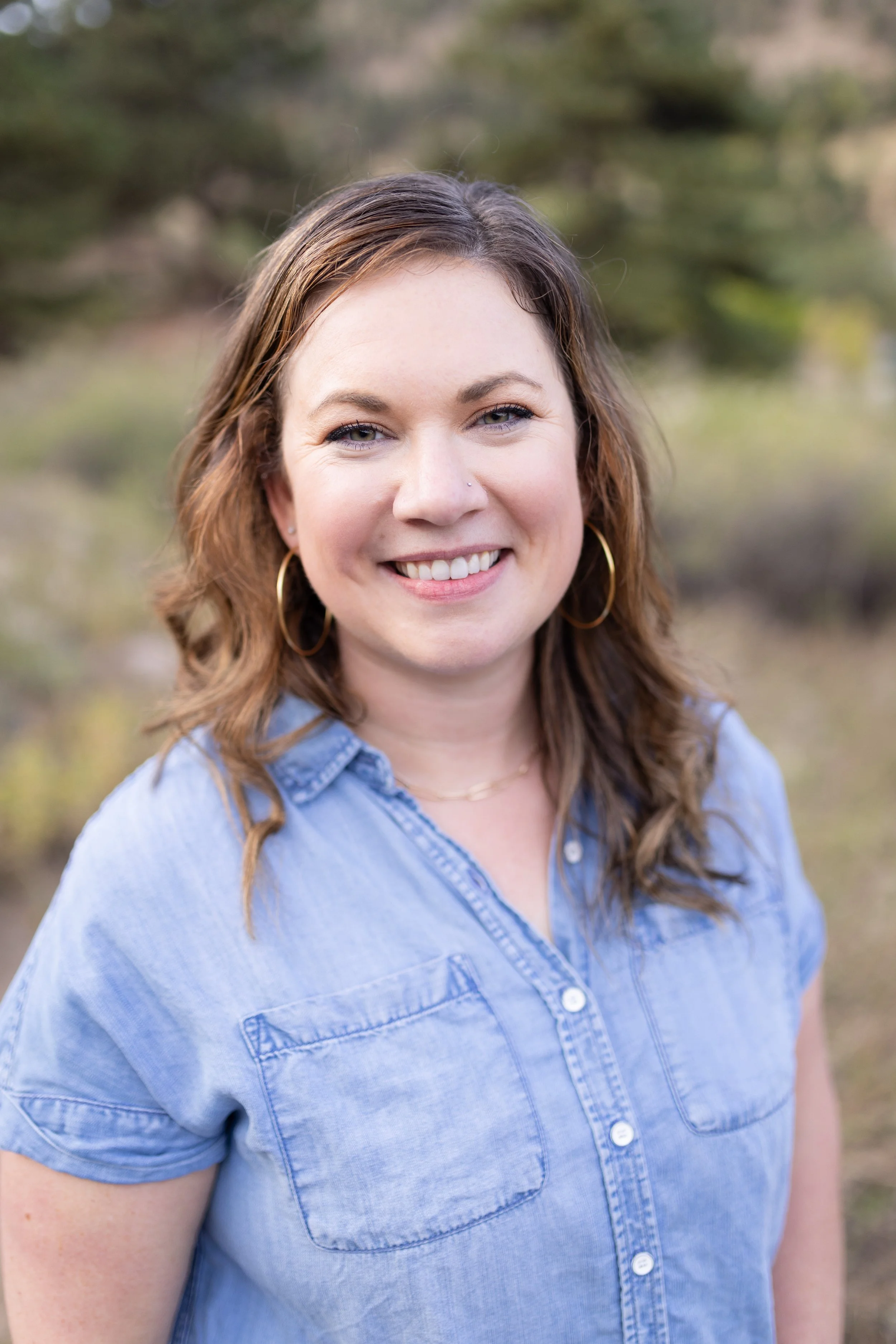 A smiling woman with brown hair, wearing a nose ring, hoop earrings, and a light blue denim shirt, standing outdoors with blurred greenery in the background.