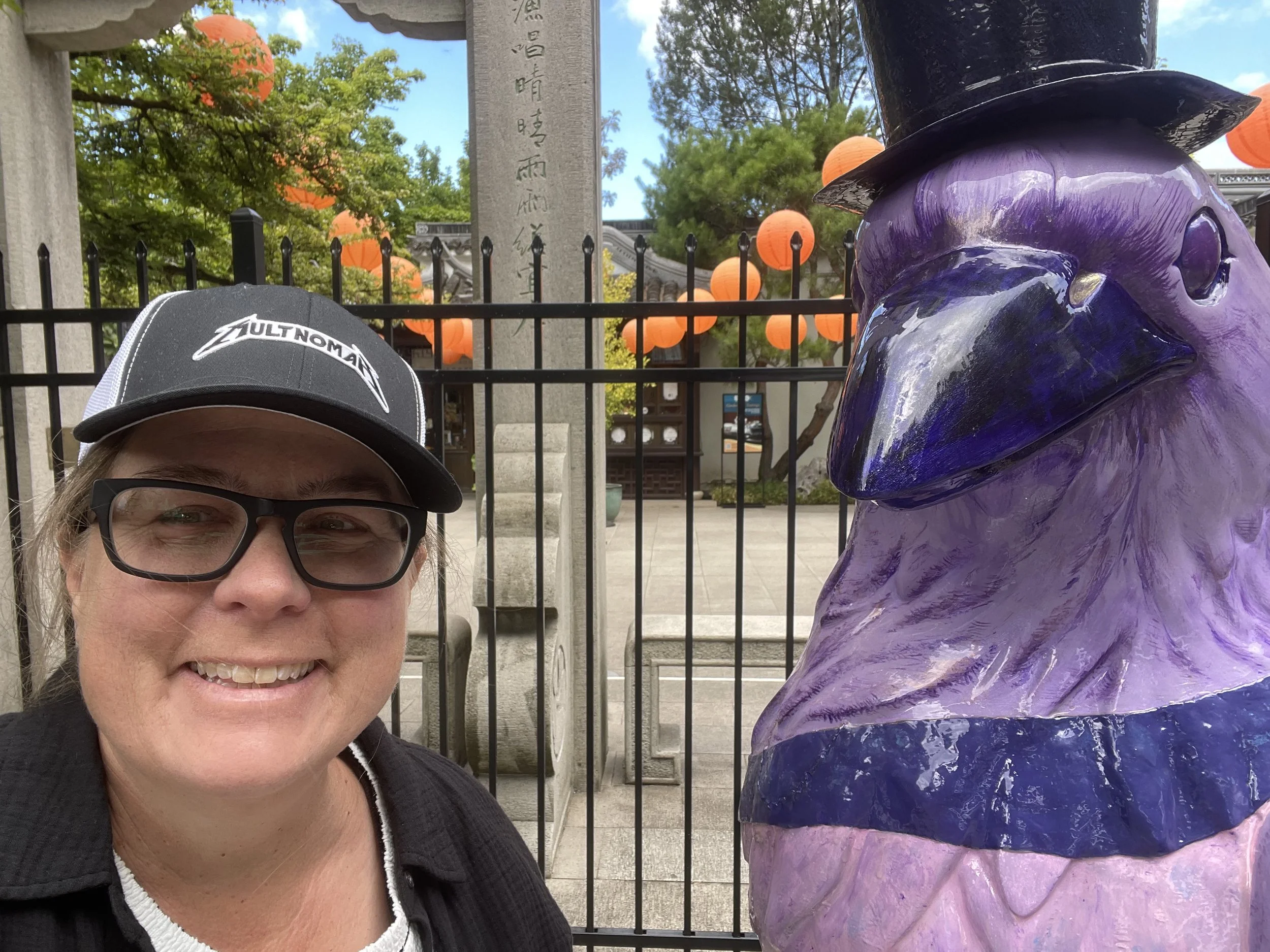 A woman with glasses and a black baseball cap takes a selfie with a purple statue of a dog wearing a black hat. The background shows trees, a stone monument with Japanese characters, and orange paper lanterns hanging from the trees.