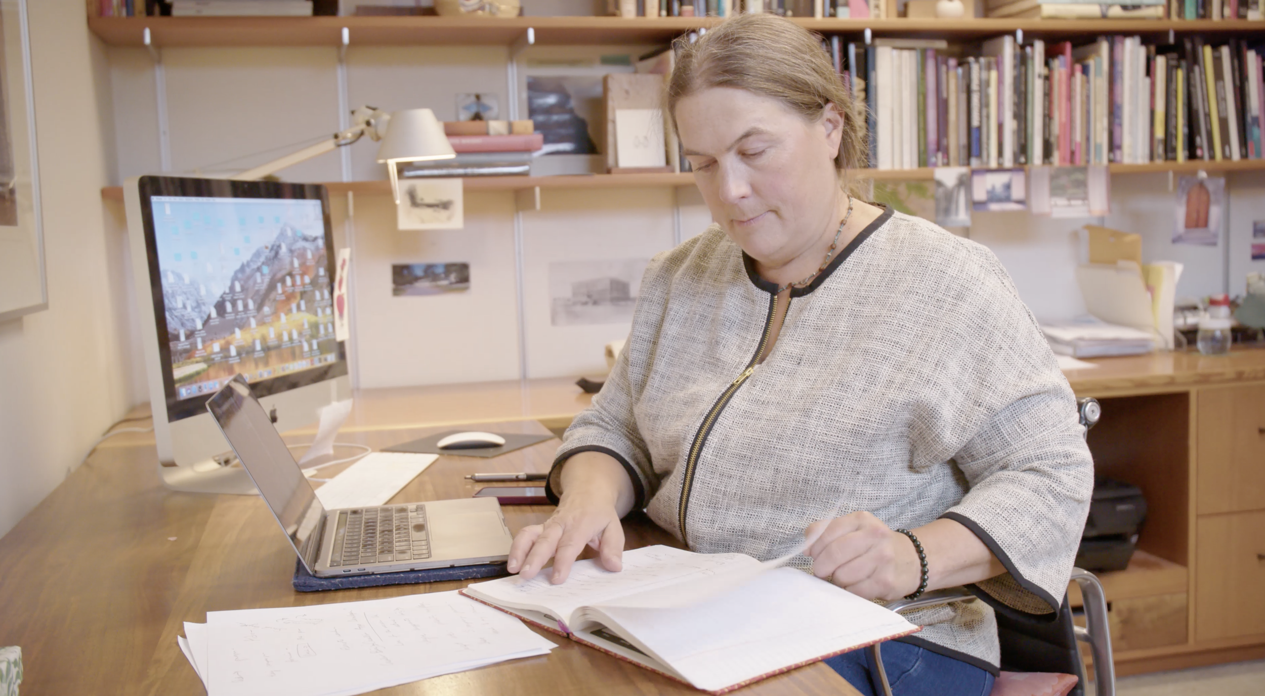 A woman with light brown hair working at a desk in an office, surrounded by papers, a laptop, and a computer monitor with a mountain desktop background. Bookshelves and photos are visible in the background.