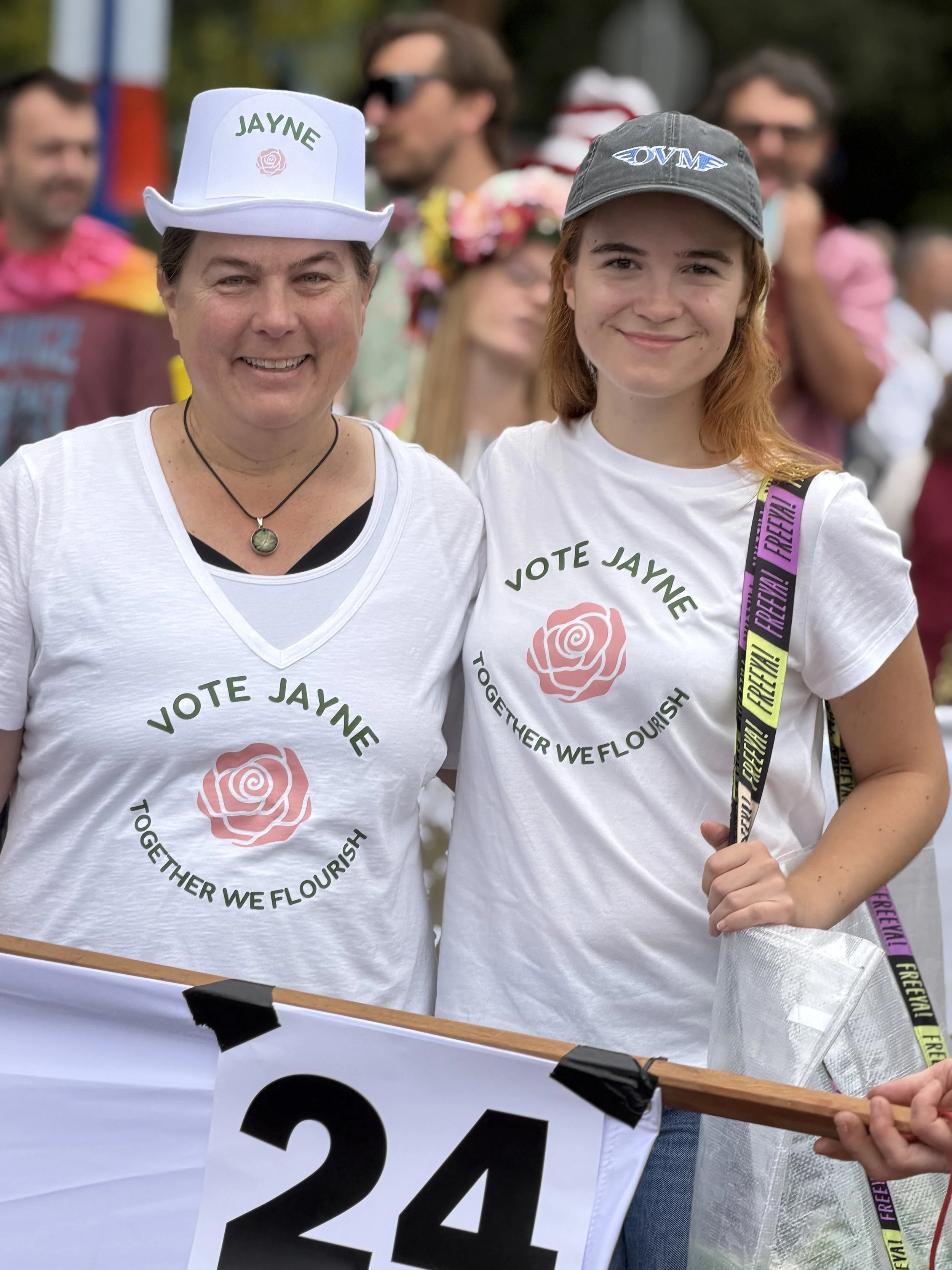 Two women at a public event wearing white T-shirts with the slogan "Vote Jayne. Together We Flourish" and a pink rose logo. The woman on the left wears a white hat with her name, Jayne, on it, and the woman on the right wears a gray cap and has a purple, yellow, and black lanyard.
