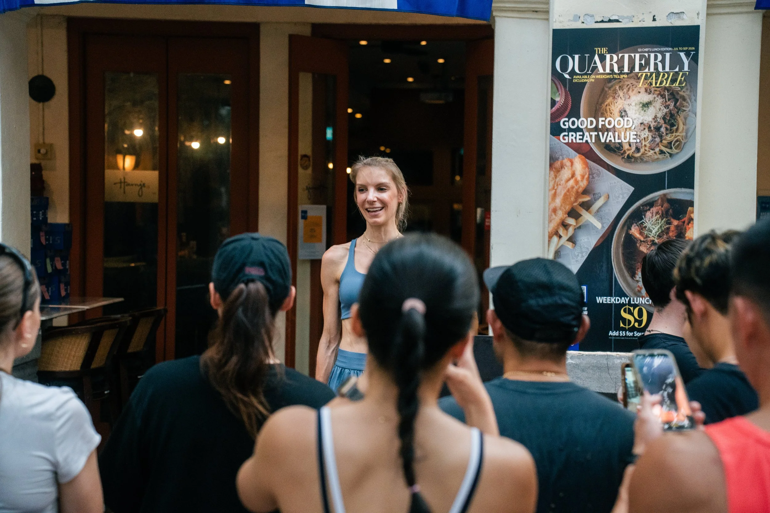 A woman wearing a blue athletic sports bra and shorts standing and smiling in front of a group of seated people in a restaurant or cafe. A large vertical poster advertising food and a lunch special is visible on the wall behind her.