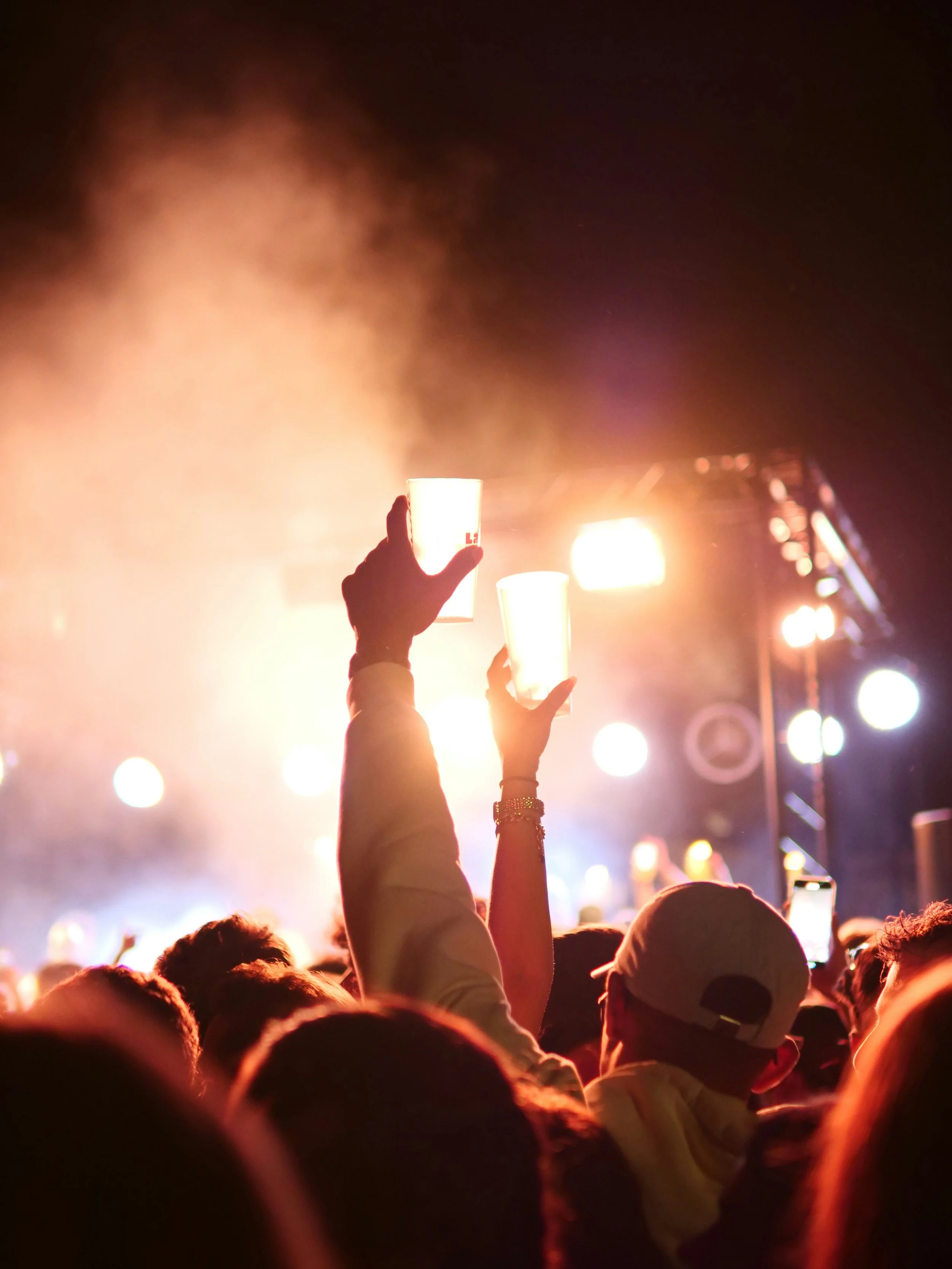People at a concert holding up illuminated cups during a performance at night, with stage lights and smoke creating a vibrant atmosphere.