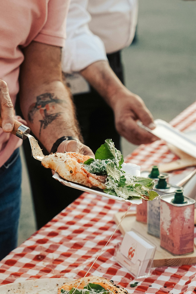 Person with tattoos holding a slice of pizza and serving it onto a plate at a food event.