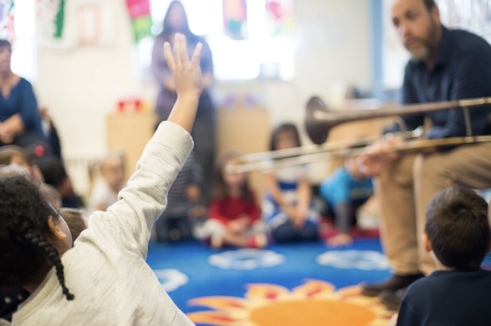 A classroom with children sitting on a colorful carpet, one child raising their hand, and a teacher playing a trombone at the front.