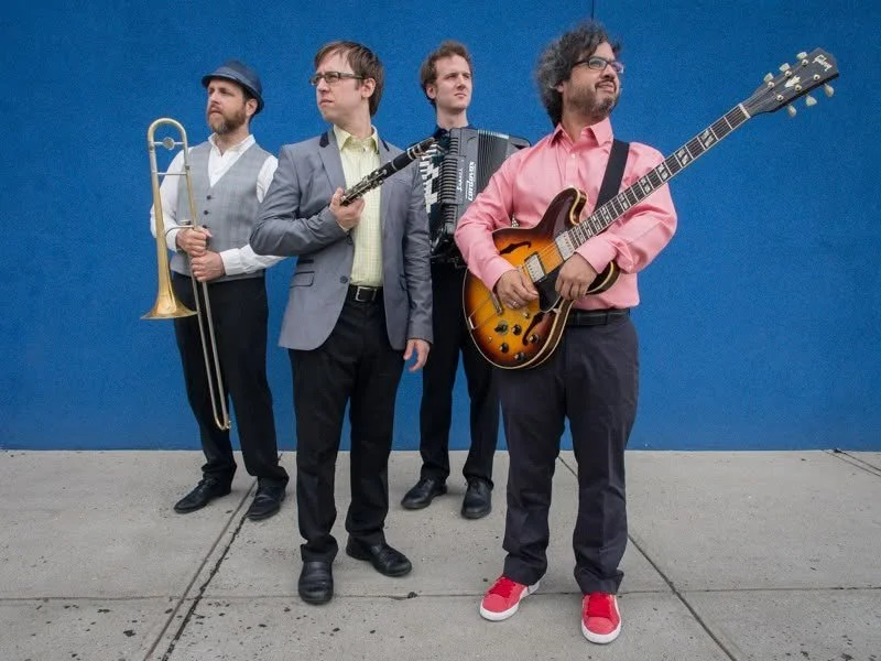 Four men standing against a blue wall, holding musical instruments, including a trombone, clarinet, accordion, and electric guitar.