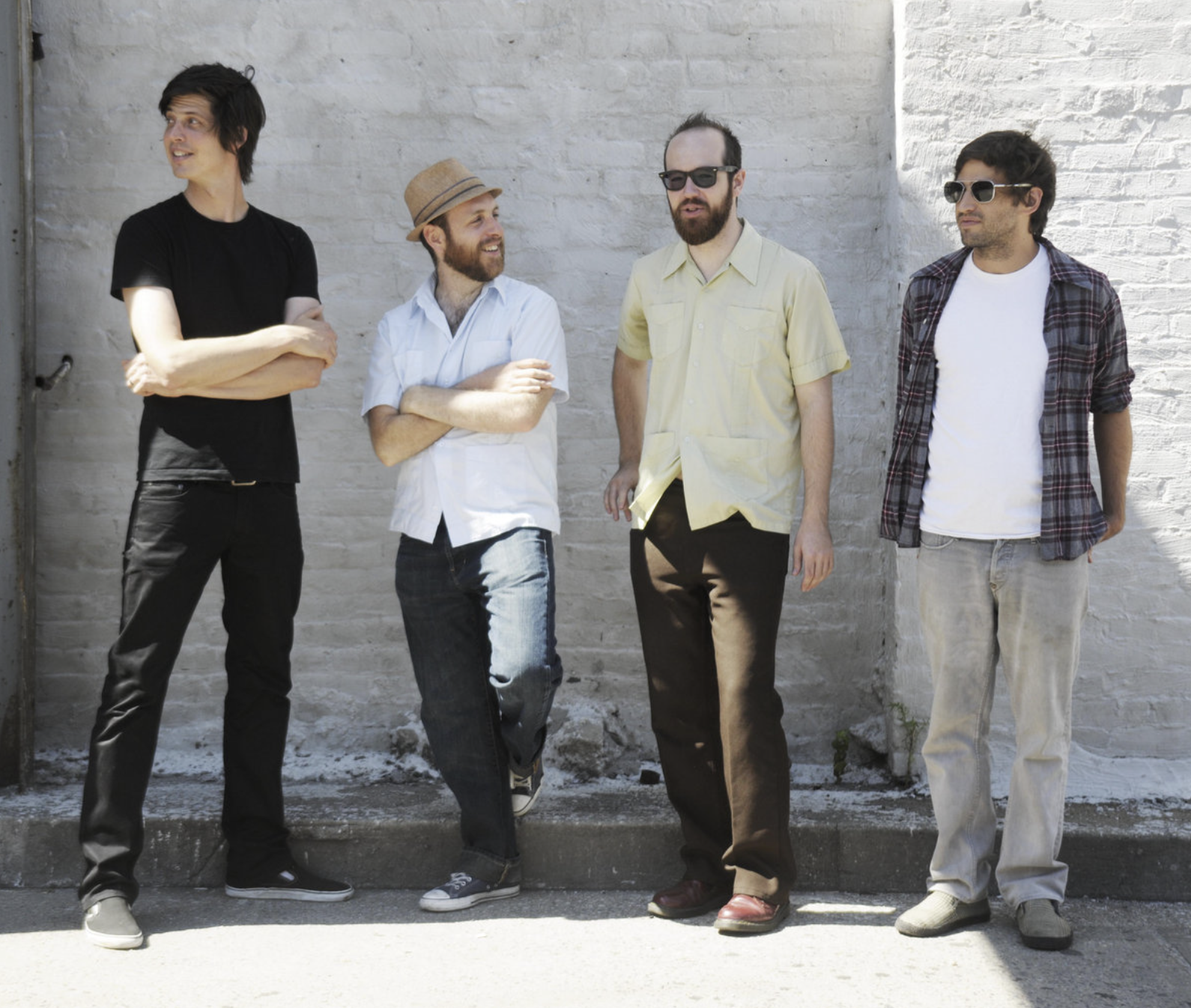 Four men standing against a white brick wall, casually dressed and engaged in conversation, some wearing sunglasses and hats.