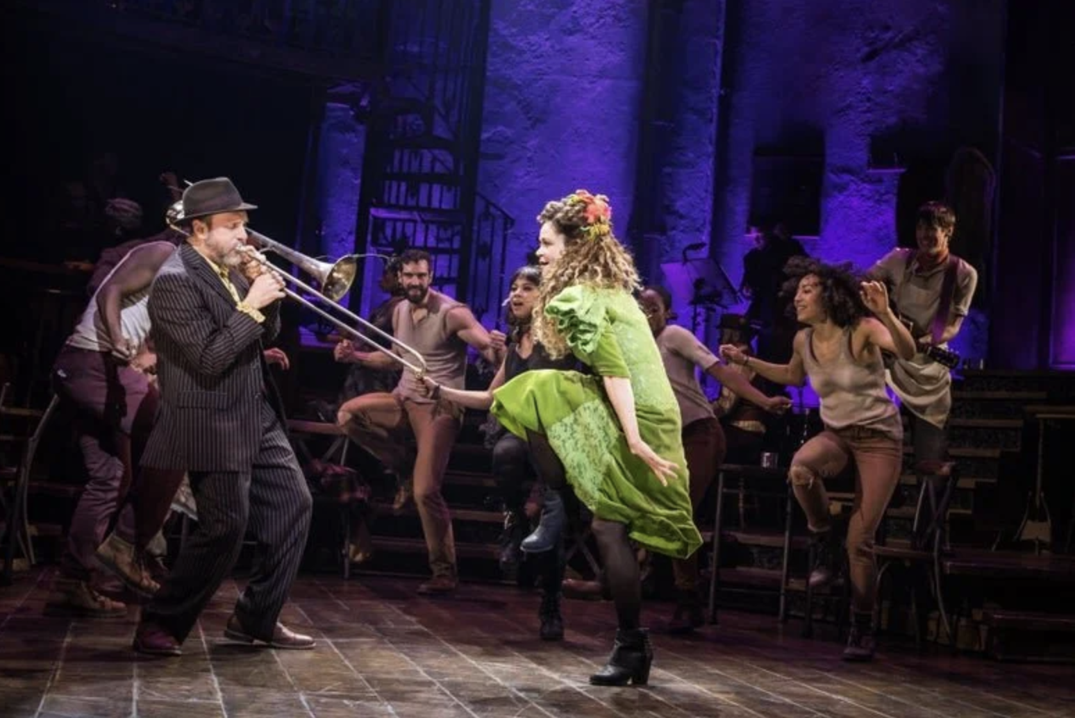 Actors on stage performing a lively musical number with a man playing trombone and a woman dancing in a green dress, surrounded by dancers, set against a vibrant purple-lit backdrop.