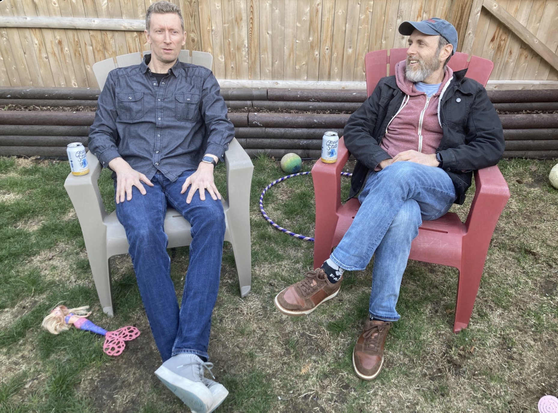 Two men sitting outdoors in plastic chairs, engaging in conversation. One man is wearing a navy blue button-up shirt, jeans, and gray sneakers, while the other is dressed in a hoodie, a jacket, jeans, and brown shoes, with a blue cap. There are cans of beer in their cups, a small toy, and a ball on the grass nearby. A wooden fence is in the background.