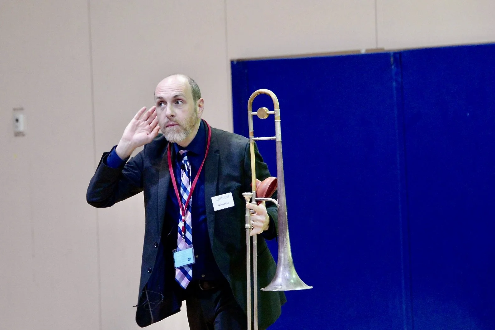 A man in a suit holds a trombone in one hand and cupped ear with the other. He is inside a room with beige walls and a blue writing surface behind him.